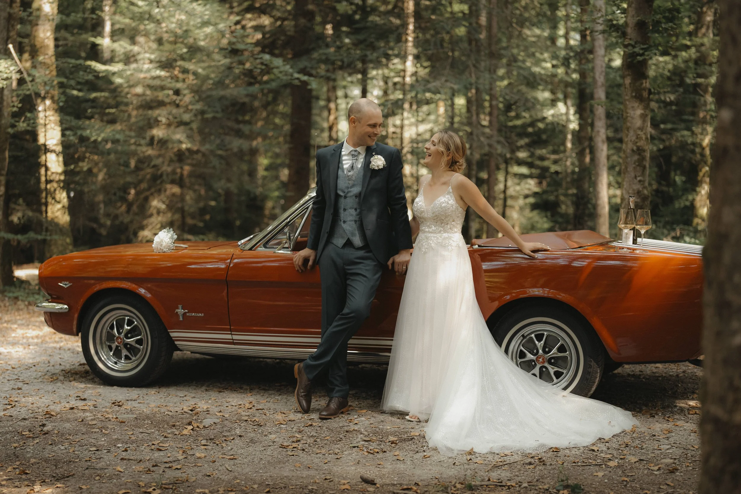 Ein Brautpaar steht in einem Wald vor einem roten Oldtimer-Cabrio, lächelnd und in Hochzeitskleidung, während sie Händchen halten.