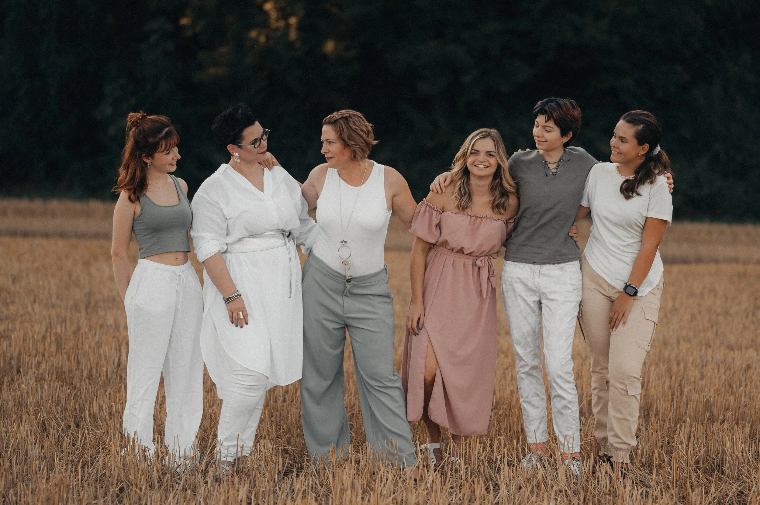 Eine Gruppe von sieben Frauen, die in einem goldenen Getreidefeld stehen, sich umarmen und lachen, bei Dämmerung.