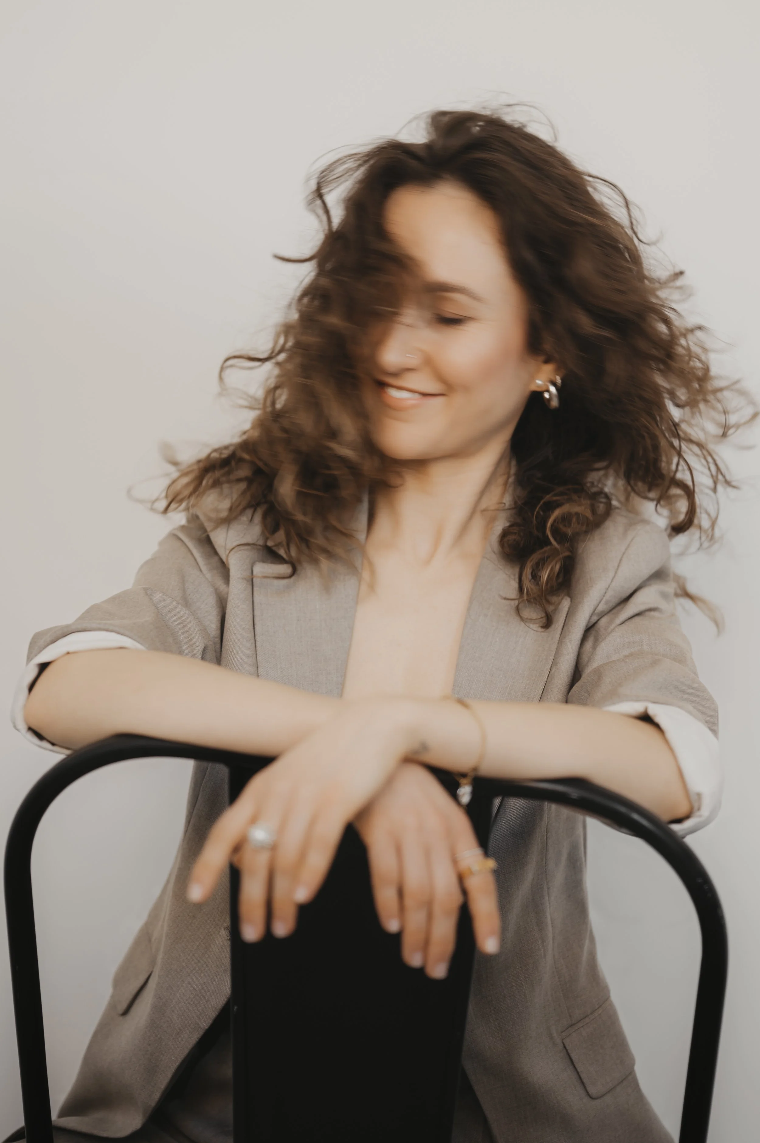Eine lachende Frau mit lockigem Haar, die auf einem schwarzen Stuhl lehnt, trägt eine graue Jacke und mehrere Schmuckstücke.