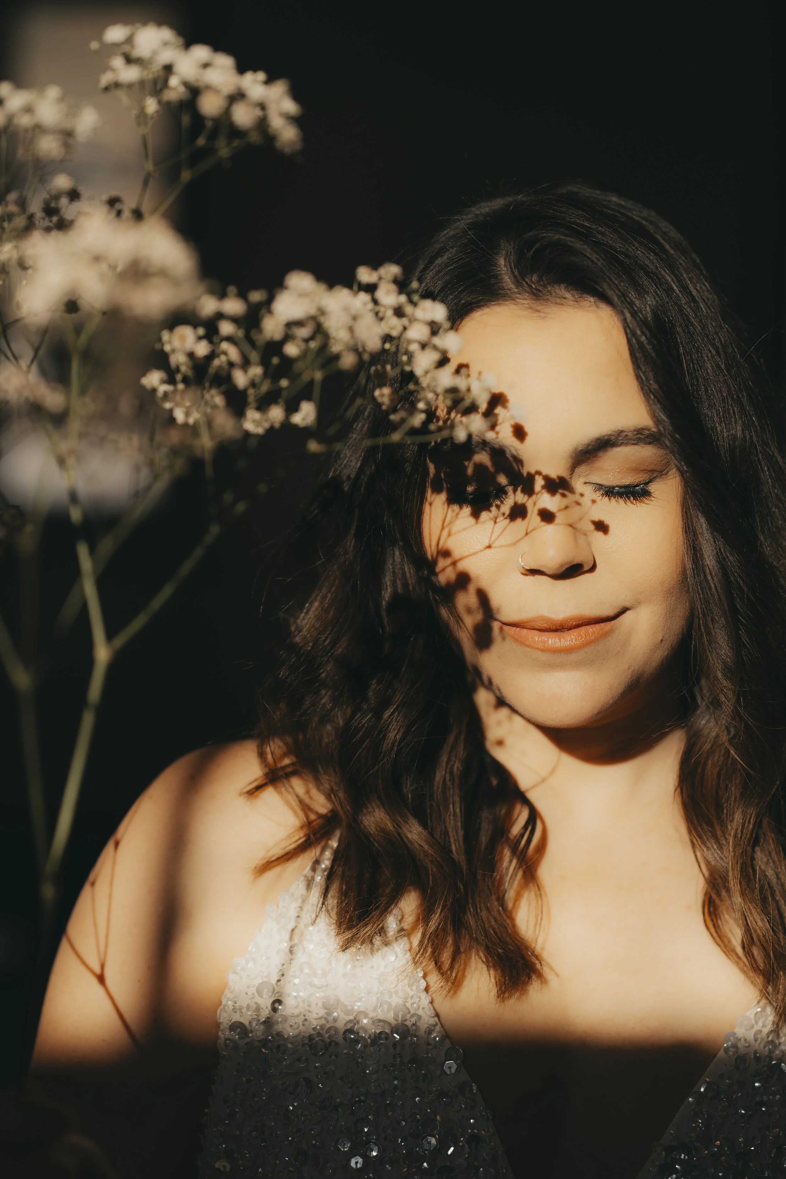 Junge Frau mit dunklen, lockigen Haaren und geschlossener Augen, trägt ein schimmerndes weißes Kleid, umgeben von Blumen, Schatten auf Gesicht und Schulter