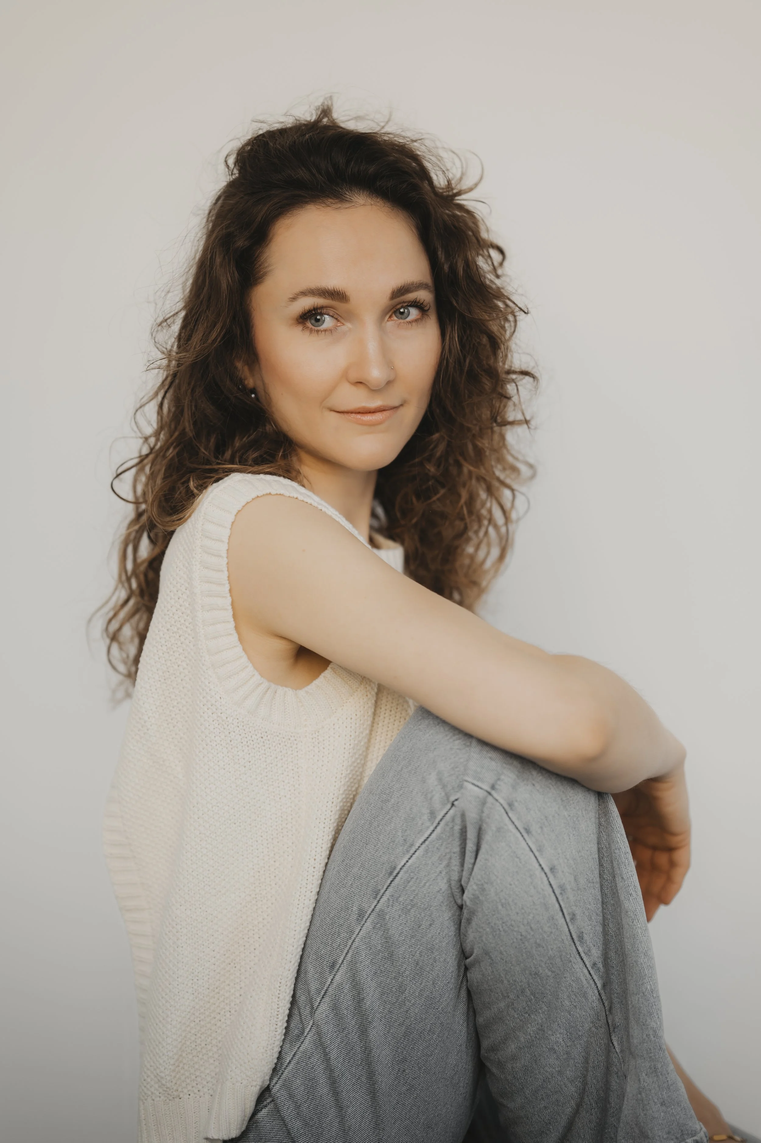 Porträt einer jungen Frau mit lockigem braunem Haar, die in die Kamera schaut, sitzend vor weißem Hintergrund.