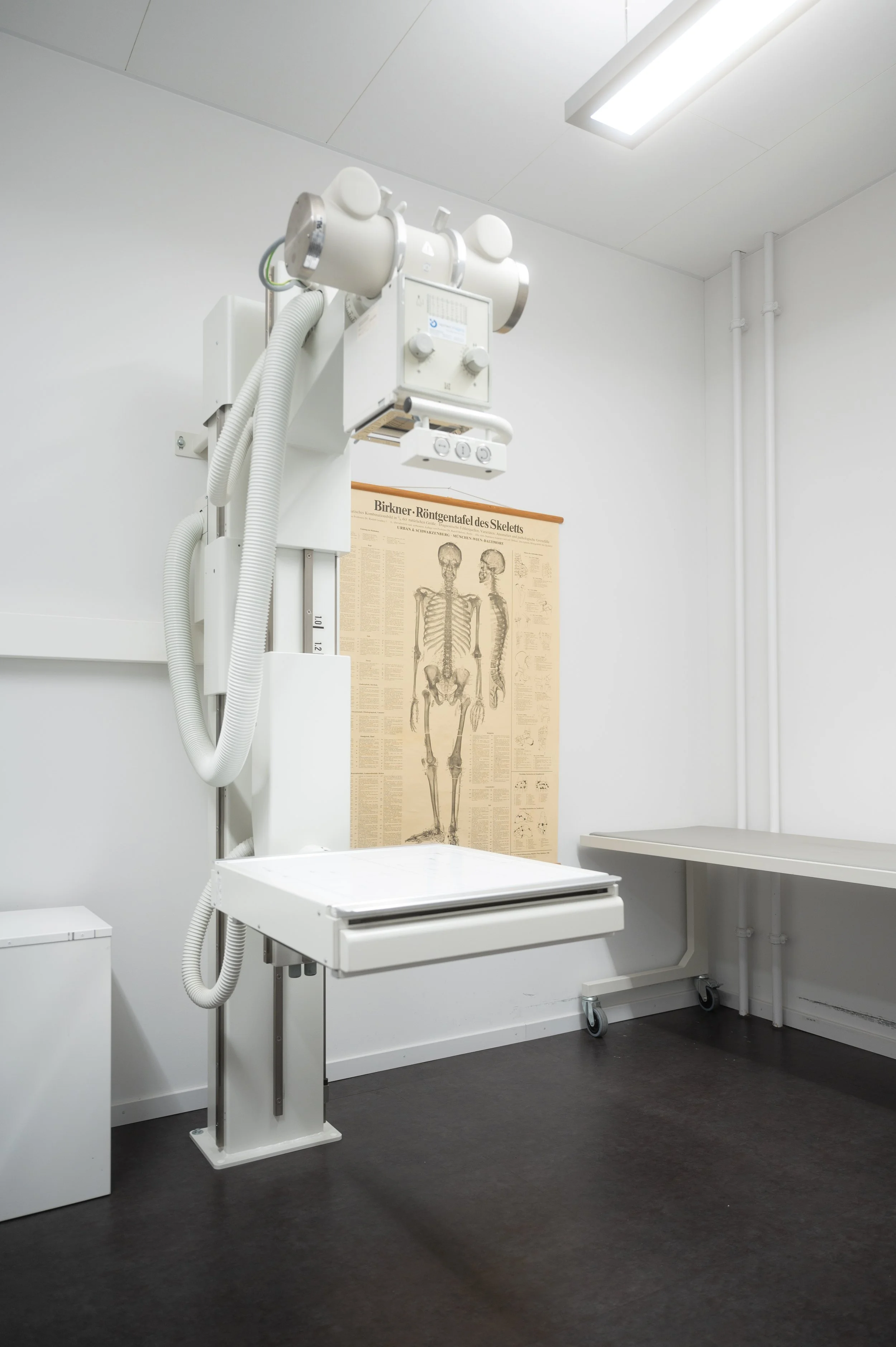 Ein Röntgengerät in einem Ärztezimmer, mit einem anatomischen Poster des menschlichen Skeletts an der Wand.