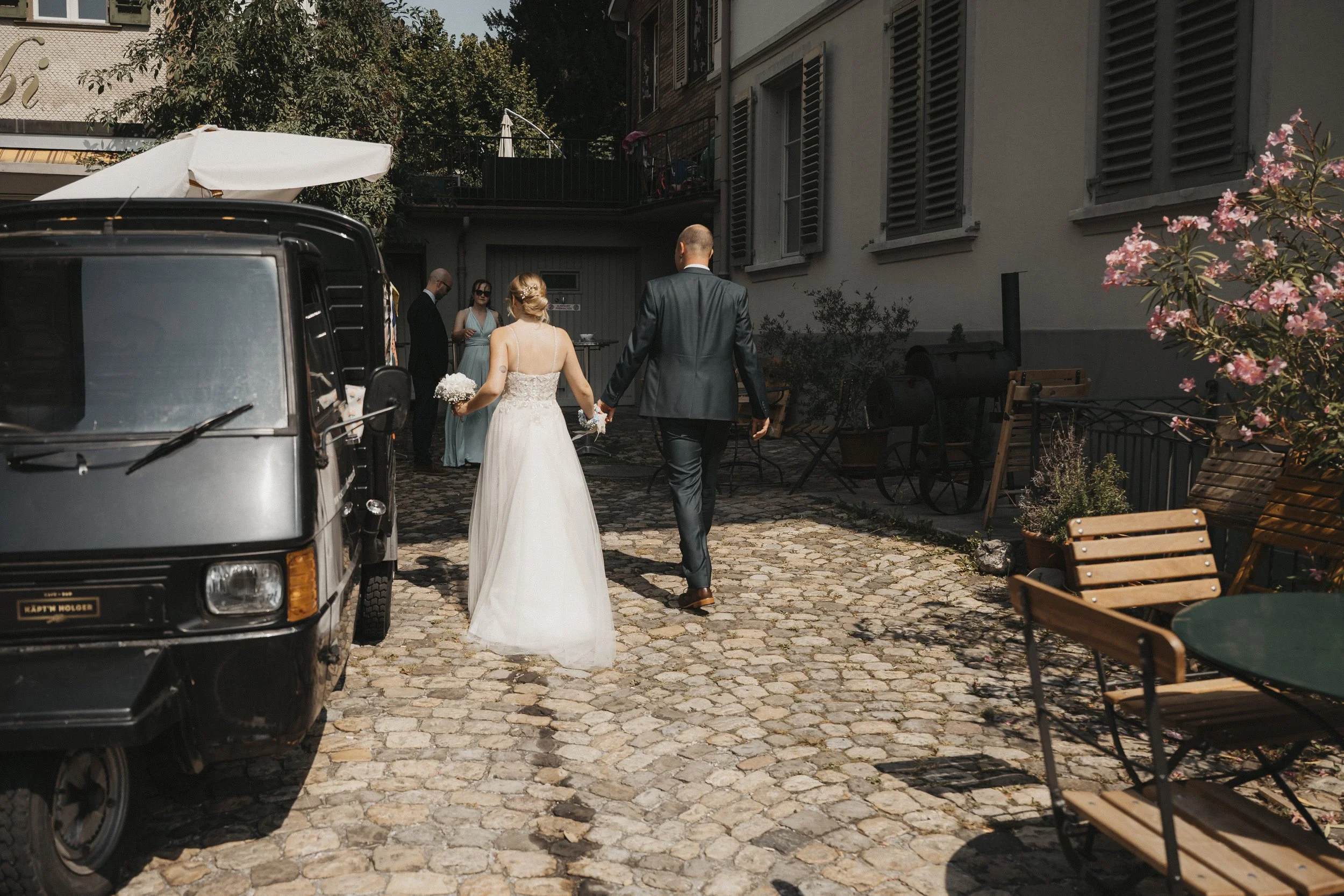 Braut und Bräutigam gehen Hand in Hand auf einer gepflasterten Straße mit Sitzgelegenheiten und Blumen, im Hintergrund sitzen Gäste bei einer Hochzeit.