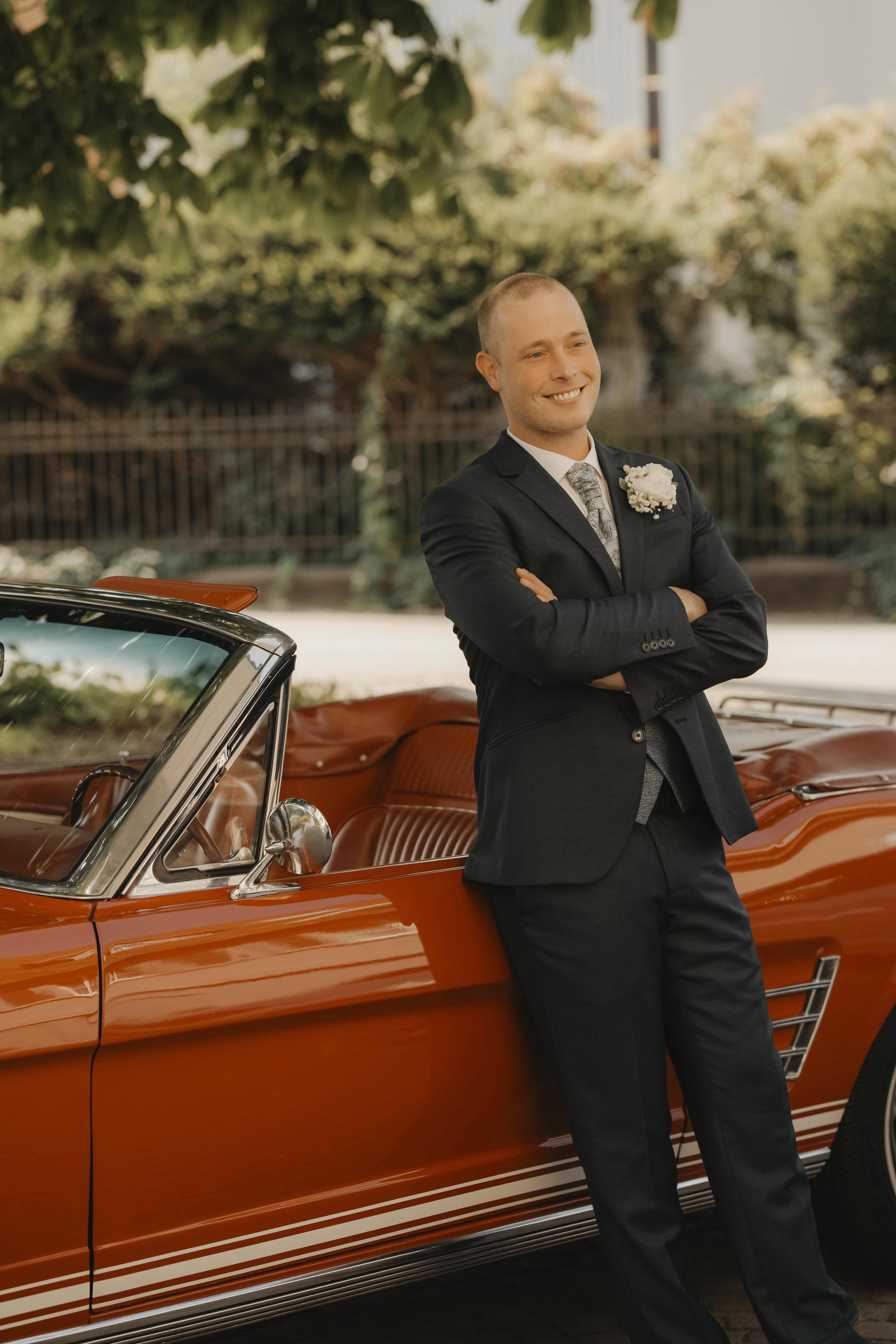 Ein Mann im Anzug lehnt an einem roten Oldtimer Cabrio, lächelt und hat die Arme verschränkt. Hintergrund mit Bäumen und einer Zaun.