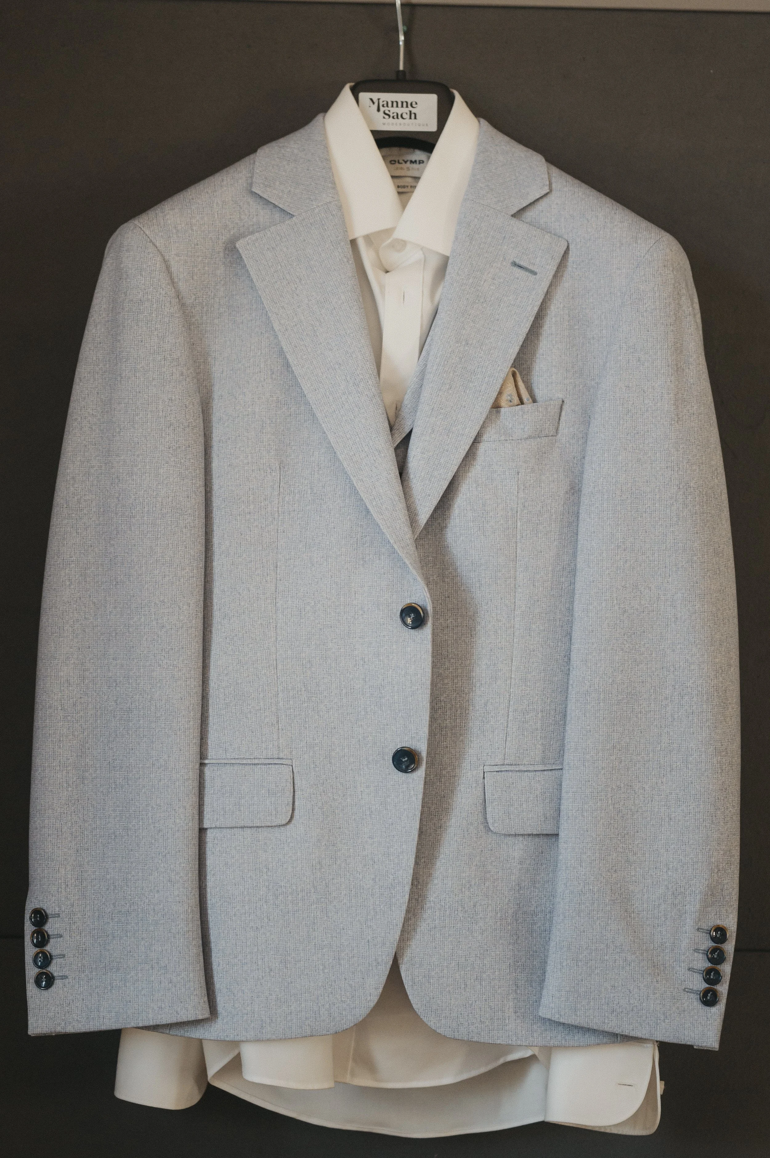 Ein hellgrauter Anzug mit weißem Hemd auf einem Hosenbügel, auf einer schwarzen Wandmontage.