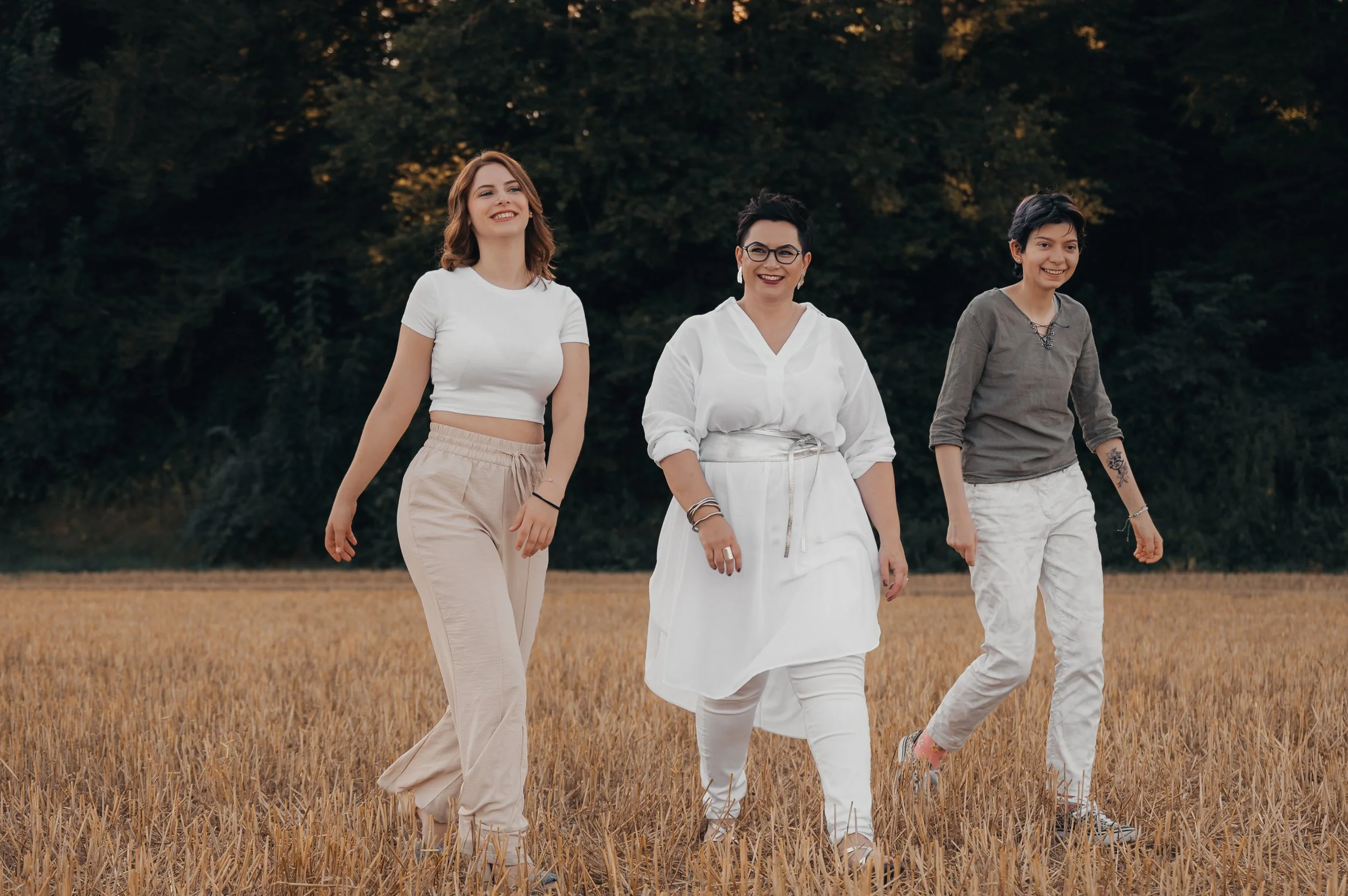 Drei Frauen gehen durch ein Getreidefeld, lachend und entspannt, im Hintergrund Bäume bei Sonnenuntergang.