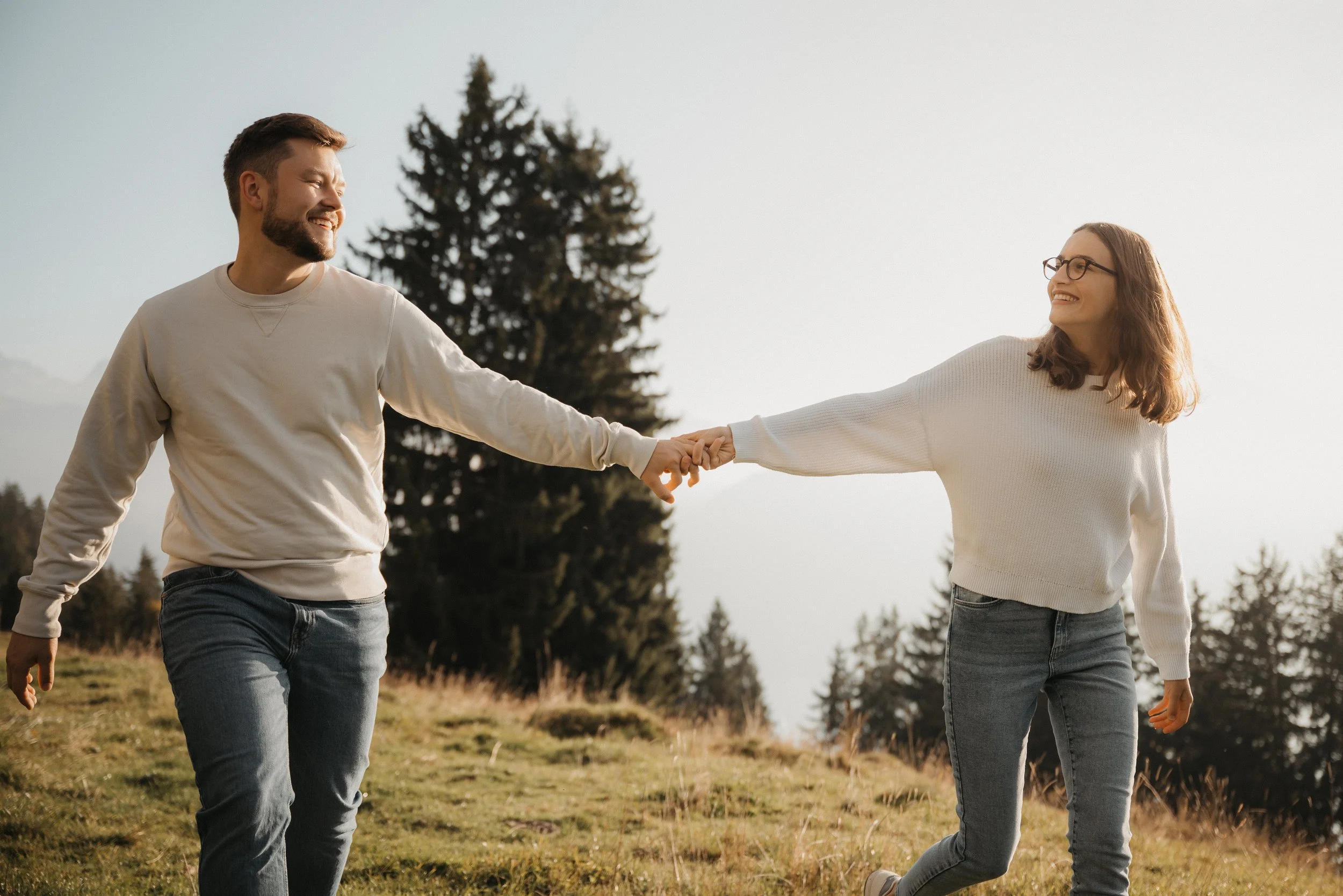 Ein Mann und eine Frau, die sich in der Natur die Hände reichen und dabei lachen, mit Bäumen im Hintergrund.