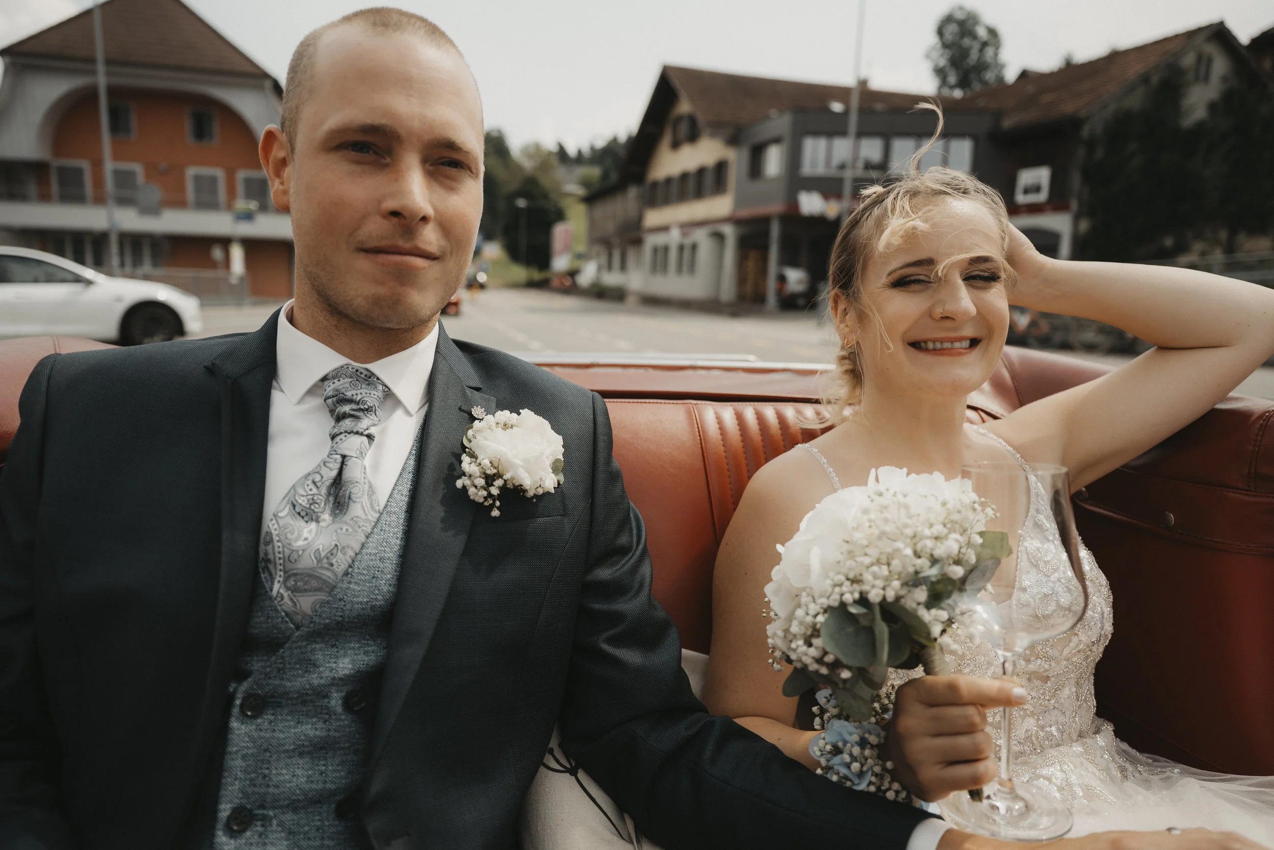 Ein frisch vermähltes Paar sitzt in einem Oldtimer, die Braut hält ein Blumenbouquet und trägt ein weißes Brautkleid, während der Bräutigam einen dunklen Anzug trägt, in einer kleinen Stadt mit traditionellen Gebäuden im Hintergrund.