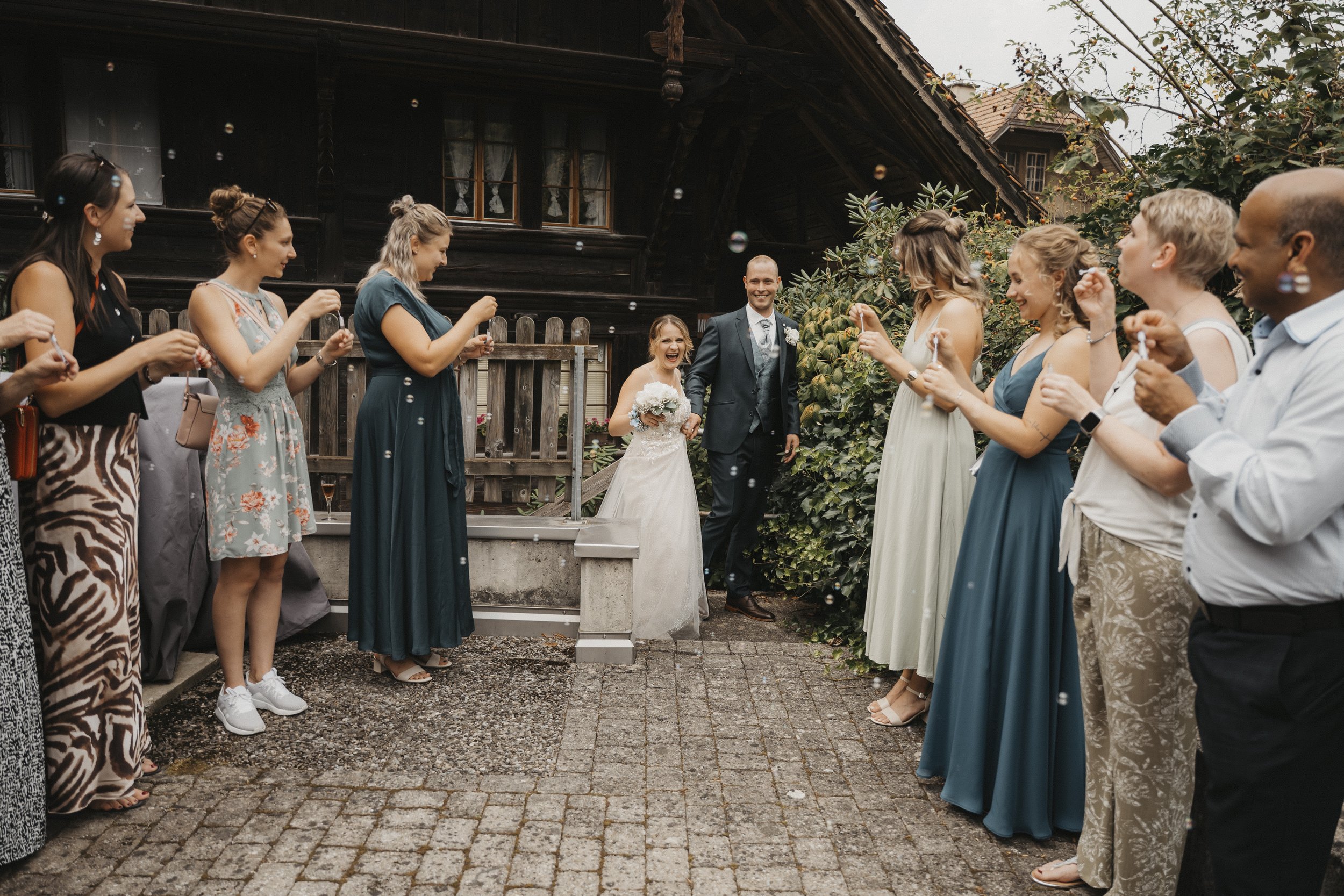 Braut und Bräutigam in der Mitte, umgeben von Freunden und Familie, die Seifenblasen machen, beim Hochzeitsempfang im Freien vor einem dunklen Holzhaus.