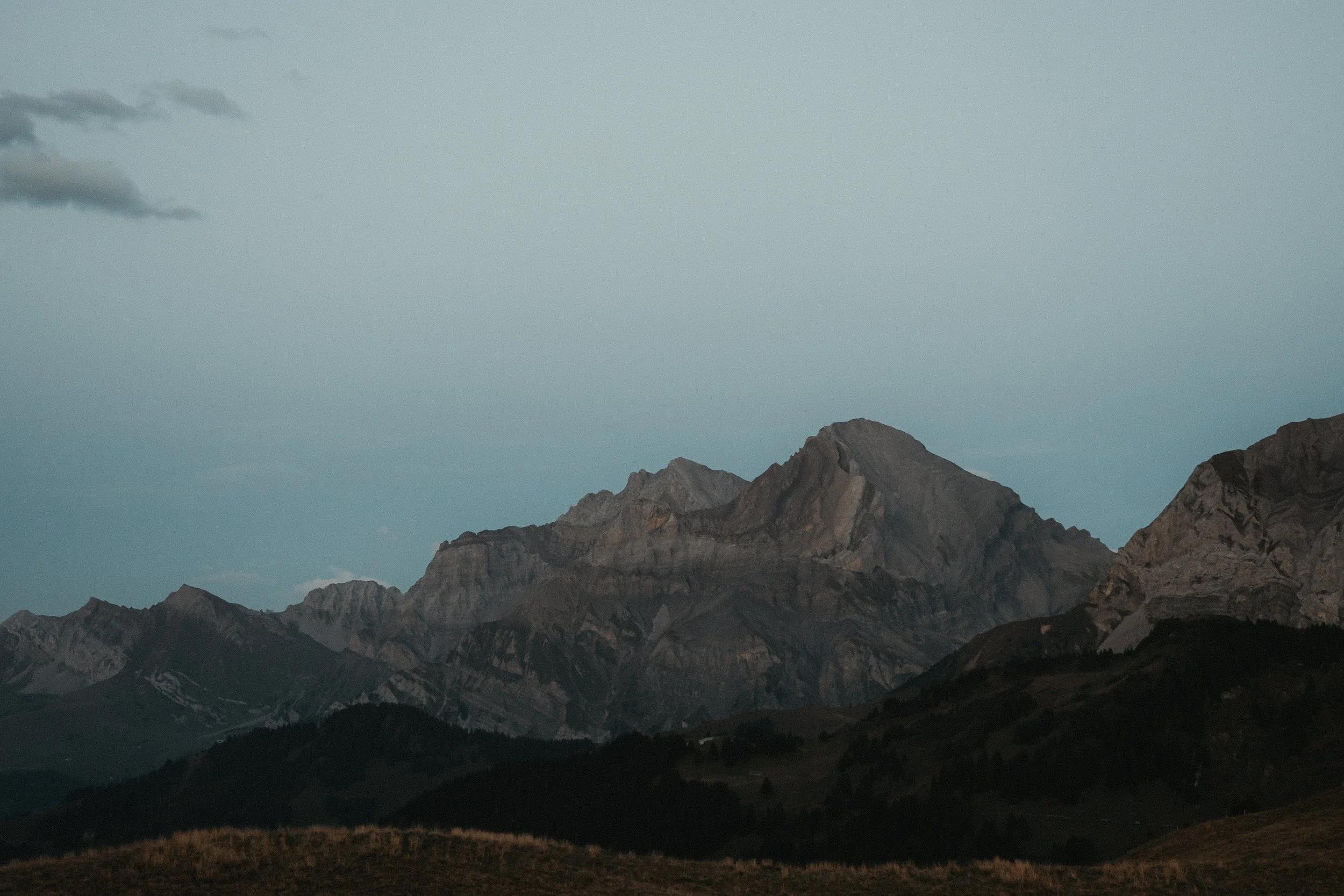 Berglandschaft mit hohen, felsigen Bergen und einer bewaldeten Hügellandschaft im Vordergrund bei Sonnenuntergang.