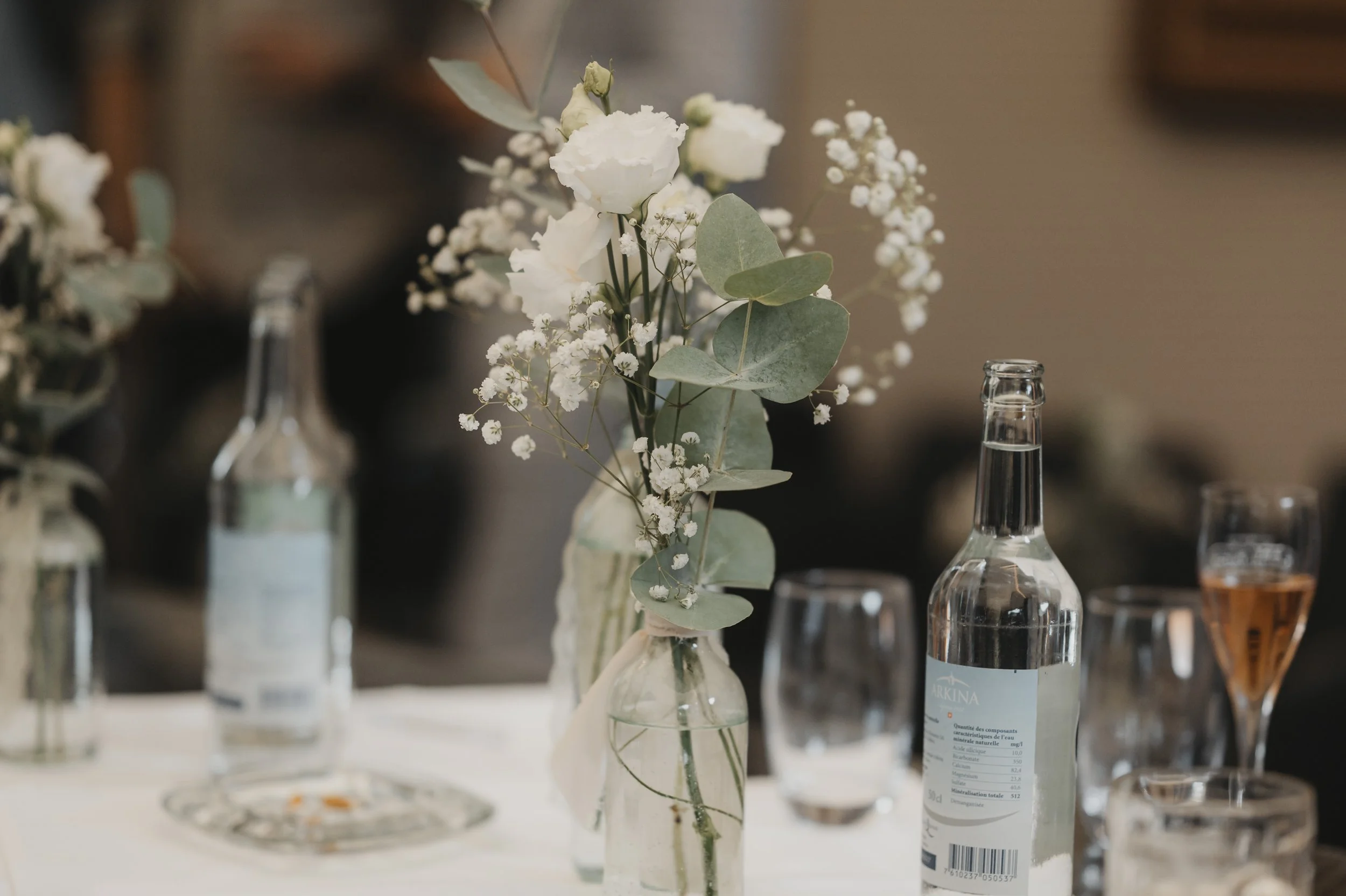 Blumenstrauß mit weißen Rosen, Eukalyptus und Gänseblümchen in einer Glasvase auf einem Tisch, umgeben von Wasserflaschen und Gläsern.