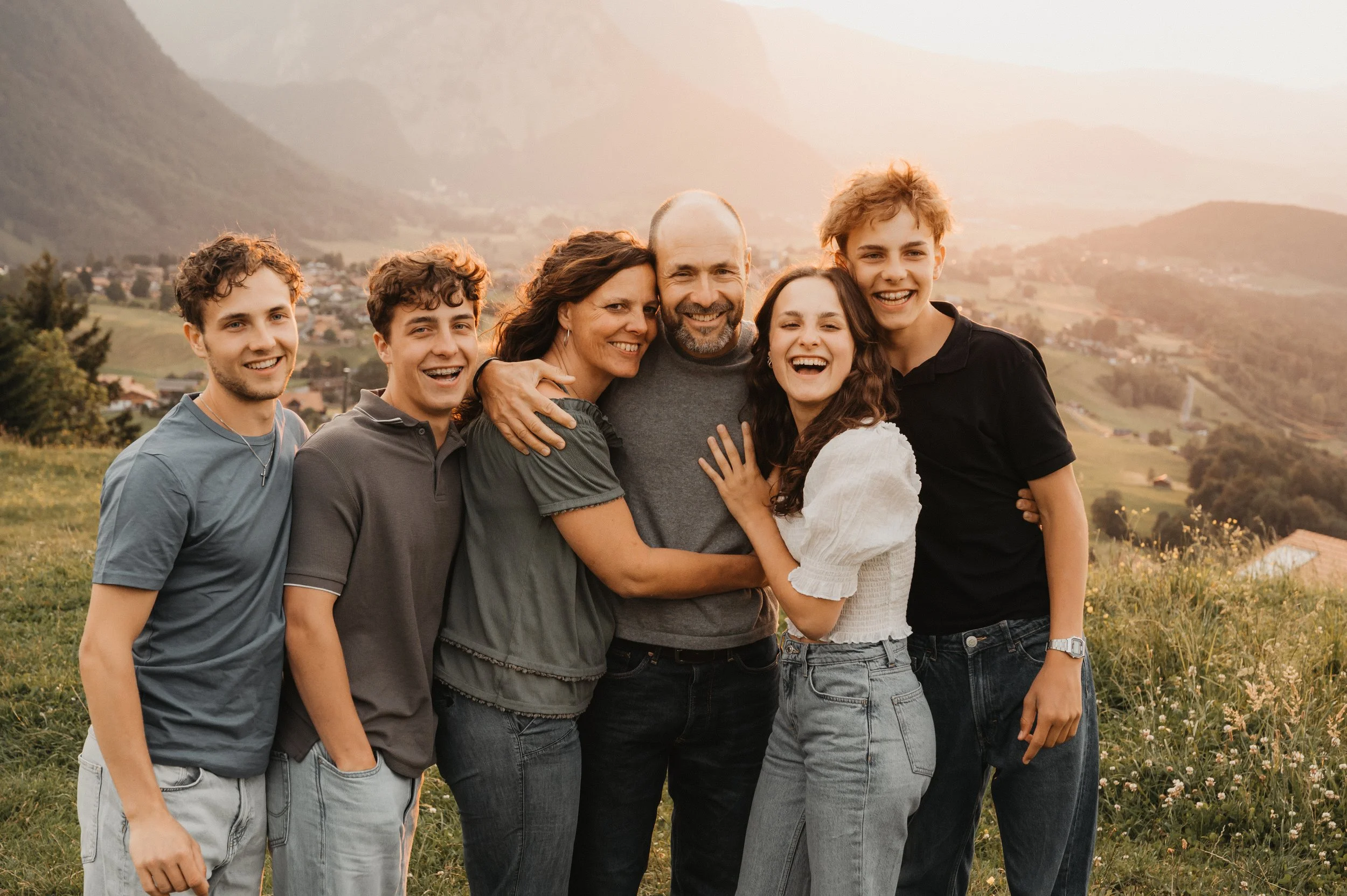 Eine Gruppe von sechs Menschen, bestehend aus zwei Erwachsenen und vier Jugendlichen, die draußen auf einer Wiese mit Bergen im Hintergrund stehen. Sie lachen und umarmen sich bei Sonnenuntergang.