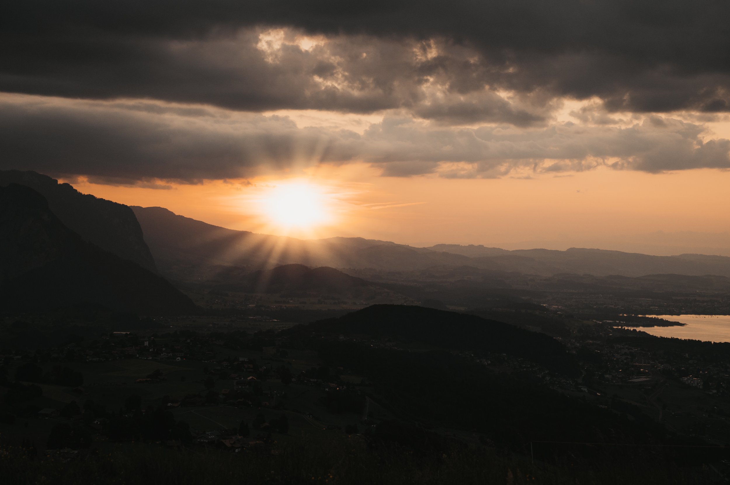 Blick auf eine Landschaft bei Sonnenuntergang mit Bergen, Wolken und einem See im Hintergrund.
