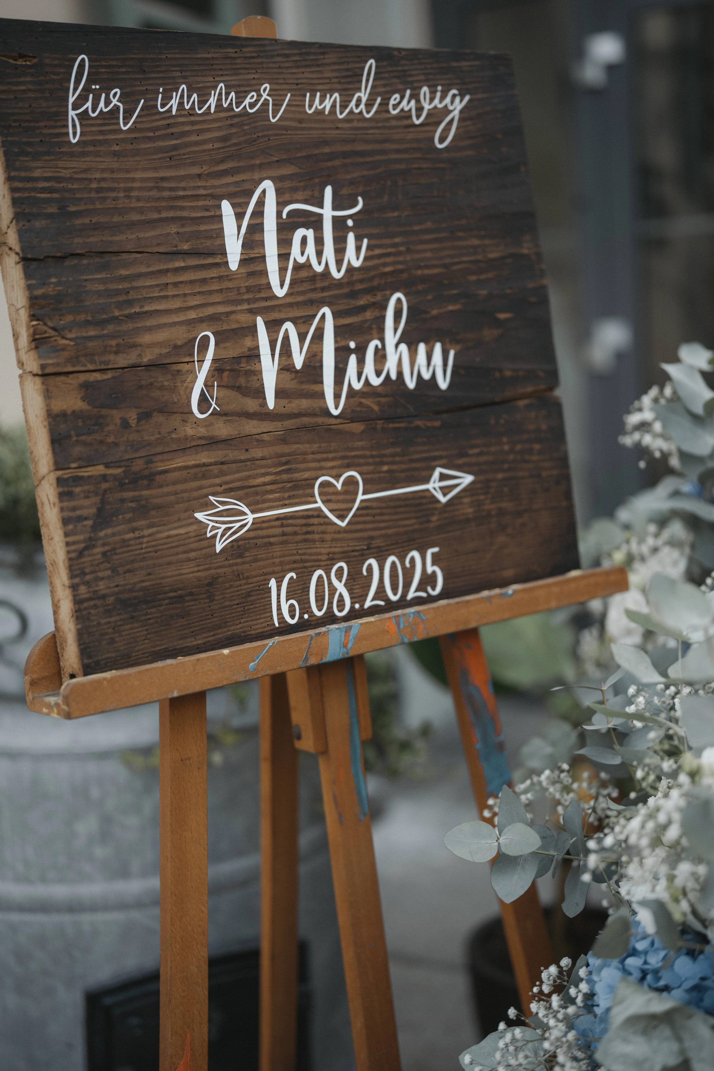 Eine Holztafel mit weißer, handgeschriebener Schrift, die besagt, dass sie für immer und ewig ist, mit den Namen Nati und Michu, einem Herz und einem Pfeil, sowie dem Datum 16.08.2025, umgeben von weißen Blumen und bläulichem Eukalyptus.