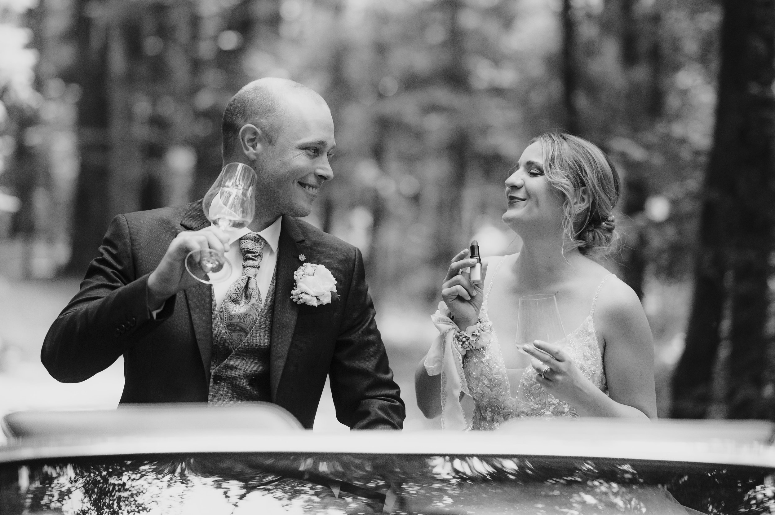 Ein Brautpaar bei einer Hochzeit im Freien, lachend und anstoßend mit Weingläsern, im Schwarz-Weiß-Foto, umgeben von Bäumen.