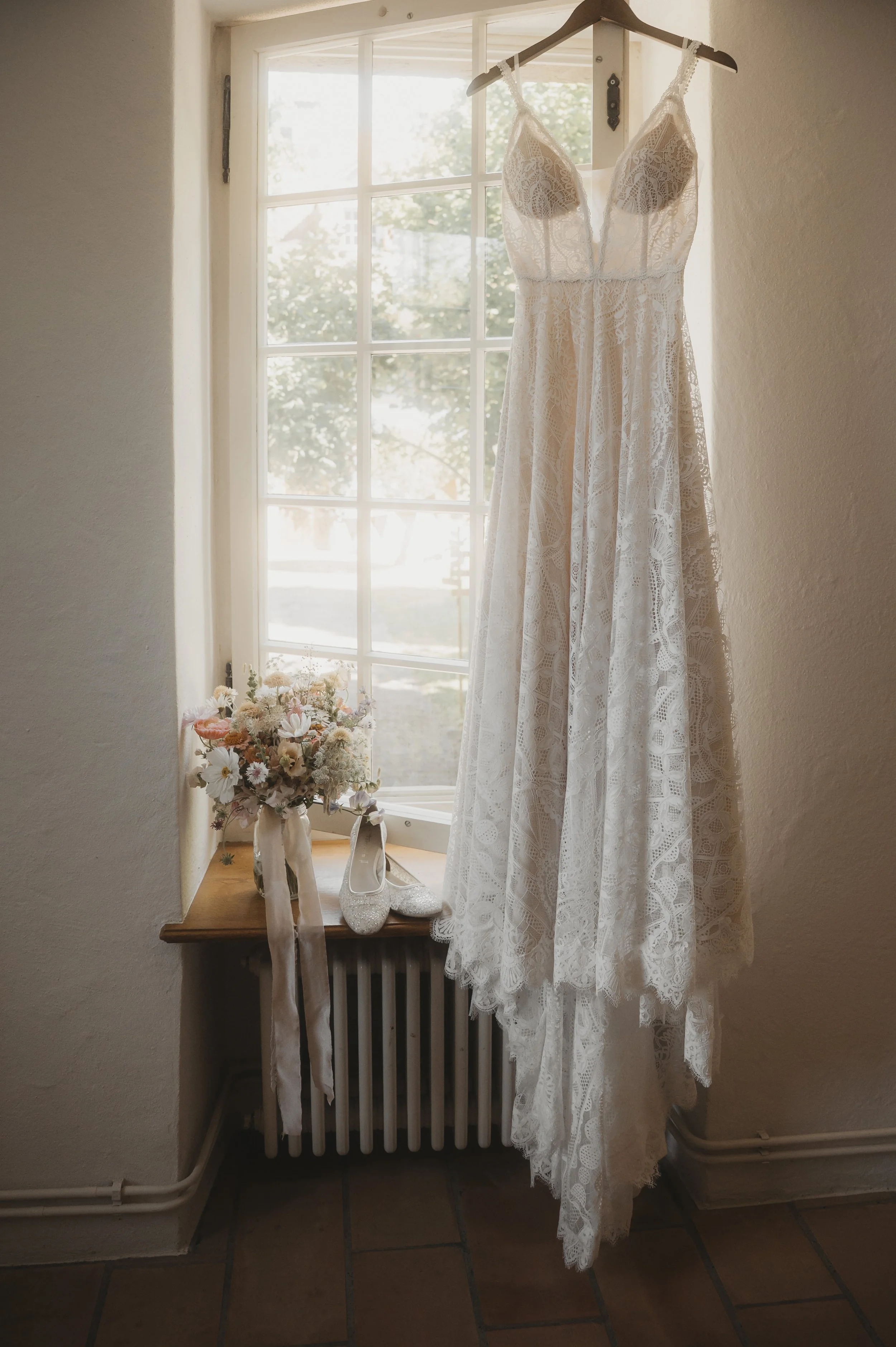 Ein weißes Brautkleid hängt an einem Kleiderbügel vor einem Fenster, darunter stehen weiße Schuhe. Neben dem Kleid liegt ein Blumenstrauß auf einem offenen Fensterbank.