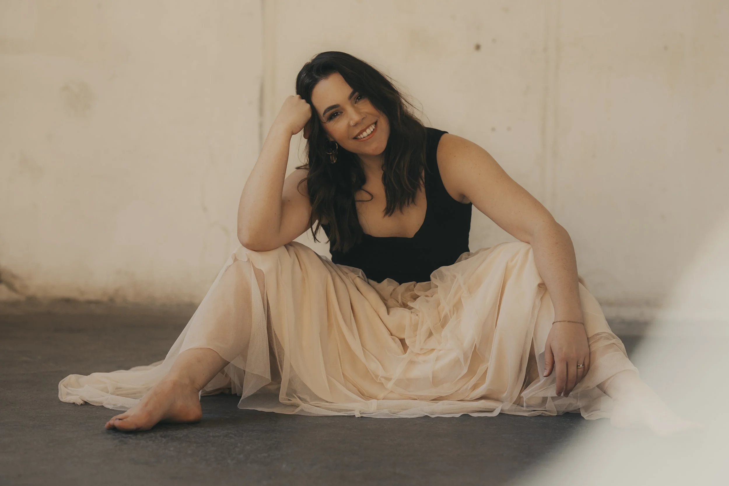 Junge Frau mit dunklen Haaren in einem schwarzen Oberteil und einem langen, pastellfarbenen Tüllrock sitzt auf dem Boden, lächelt und schaut freundlich in die Kamera.