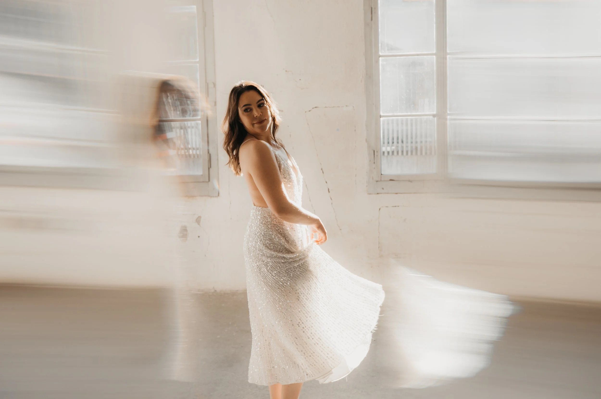 Frau in einem weißen, funkelnden Kleid steht in einem hellen Raum mit großen Fenstern, die Sonnenlicht hereinlassen.