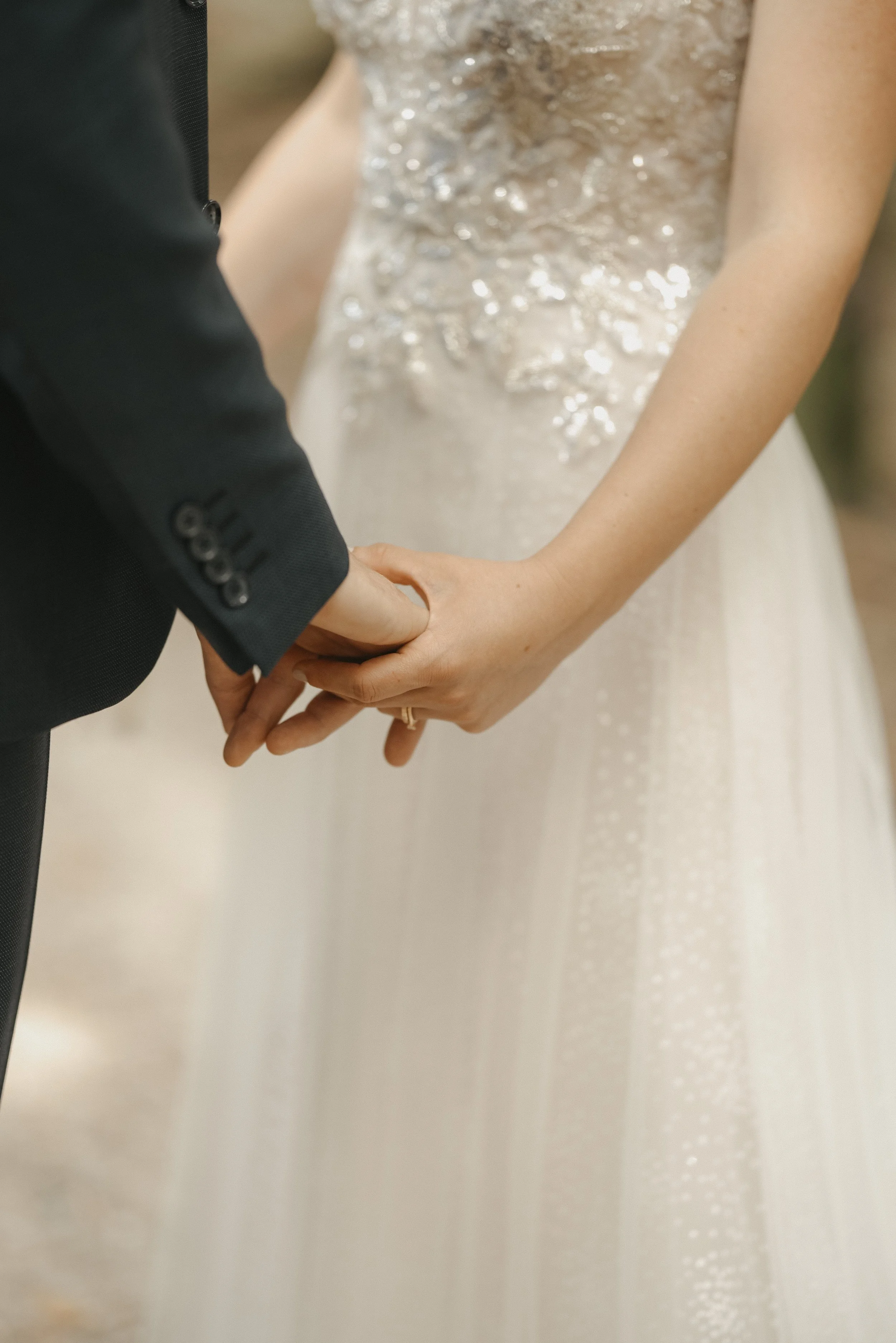 Ein Brautpaar hält sich an den Händen während ihrer Hochzeit. Der Fokus liegt auf den Händen, einem Brautkleid mit Spitze und einem dunklen Anzug.