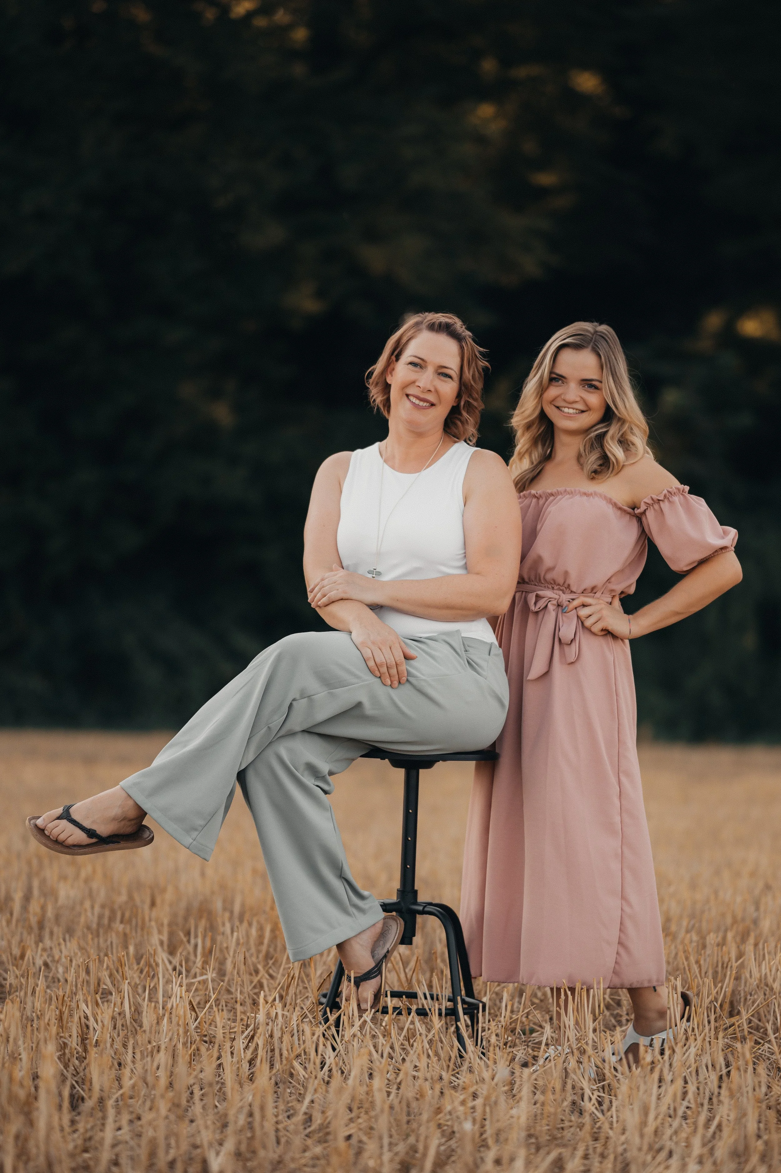 Zwei Frauen stehen und sitzen in einem Feld, die ältere sitzt auf einem Stuhl, die jüngere steht neben ihr, beide lächeln und schauen in die Kamera. Im Hintergrund sind Bäume.