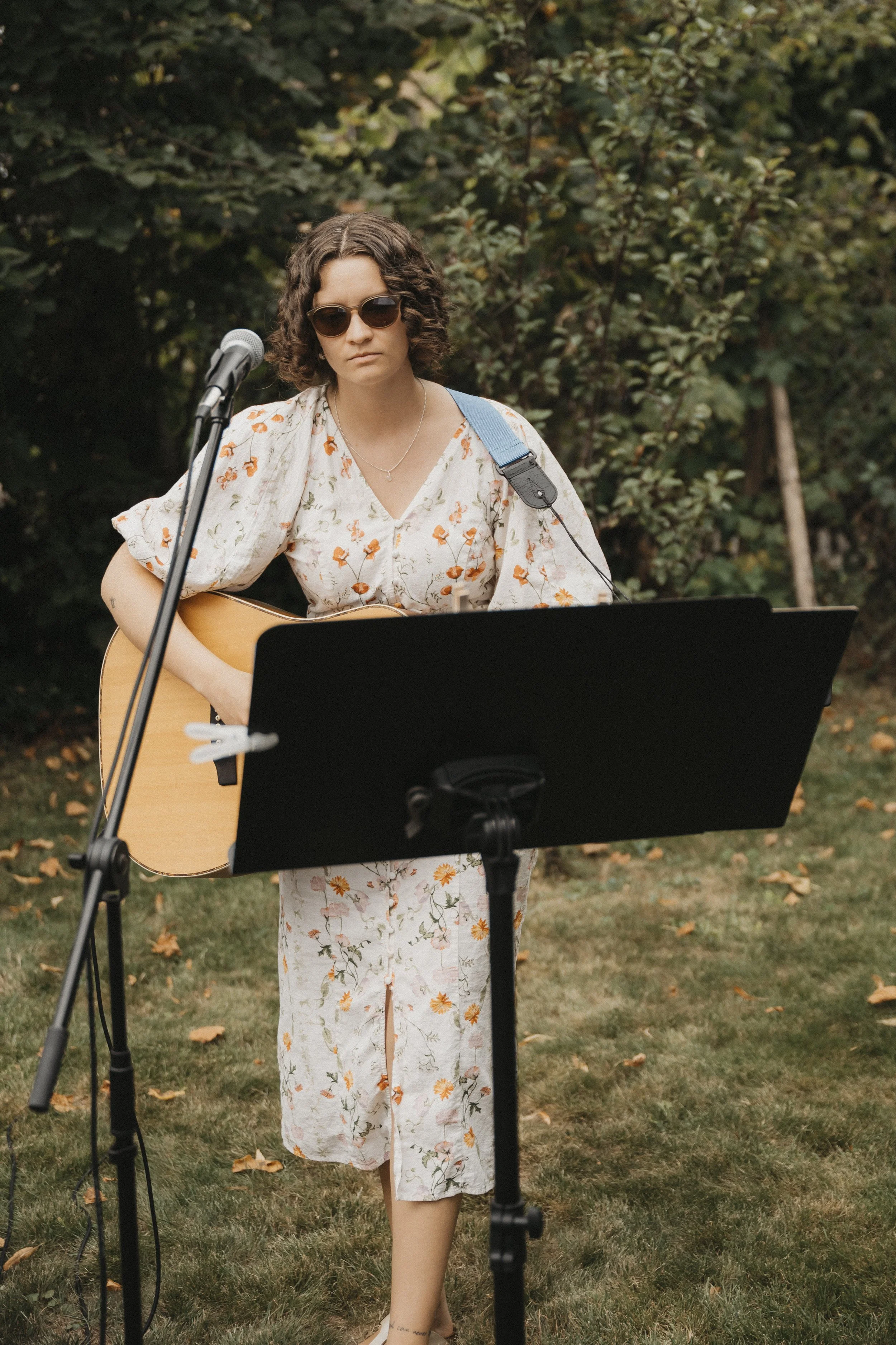 Junge Frau mit dunklen runden Sonnenbrillen, gebleichtem Haar und heller Baumwollkleidung mit Blumenmuster spielt Gitarre im Freien, vor einem Musikständer, im Garten mit Büschen und Bäumen im Hintergrund.