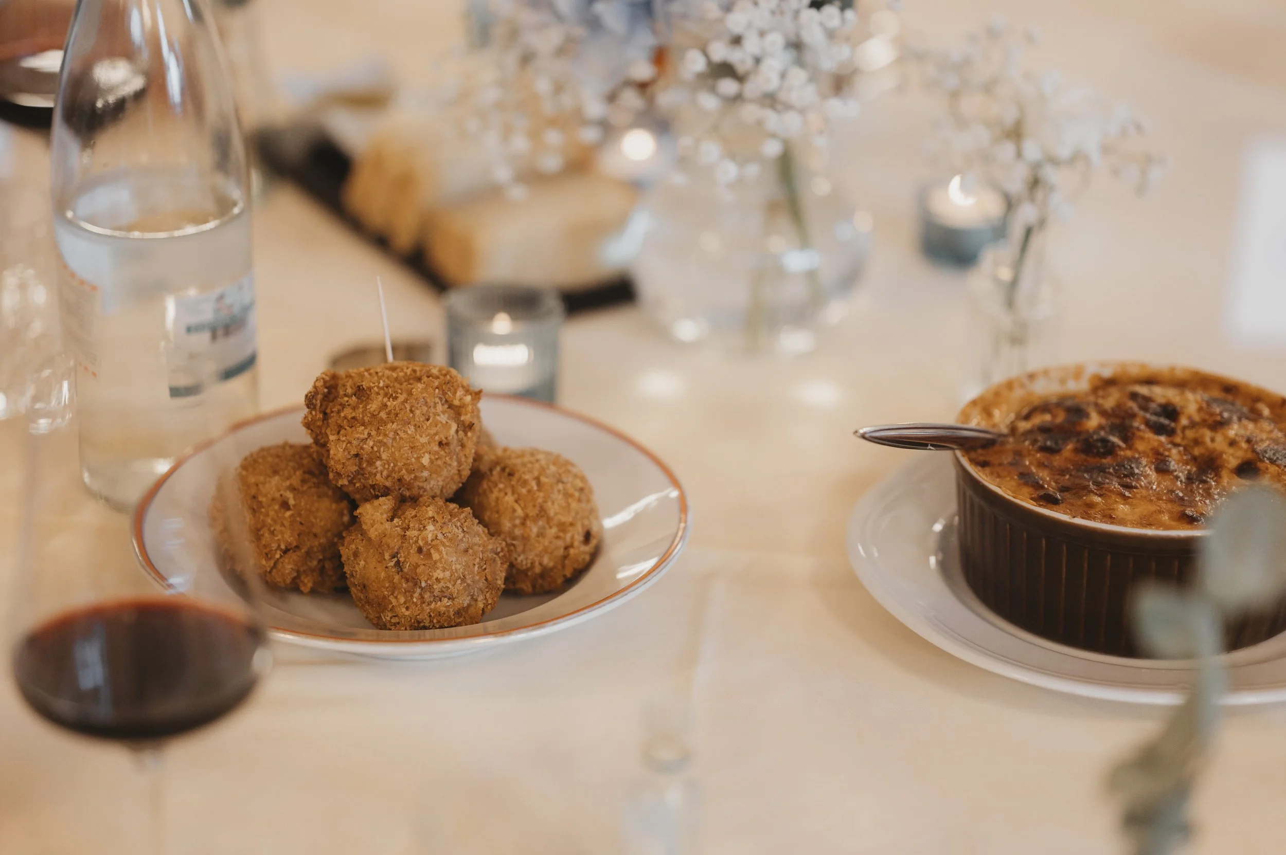 Auf einem Esstisch sind eine Schale mit frittierten Bällchen, ein Glas Wasser, ein Glas Rotwein, eine Ofenkasserolle mit Auflauf, Blumen und Kerzen.