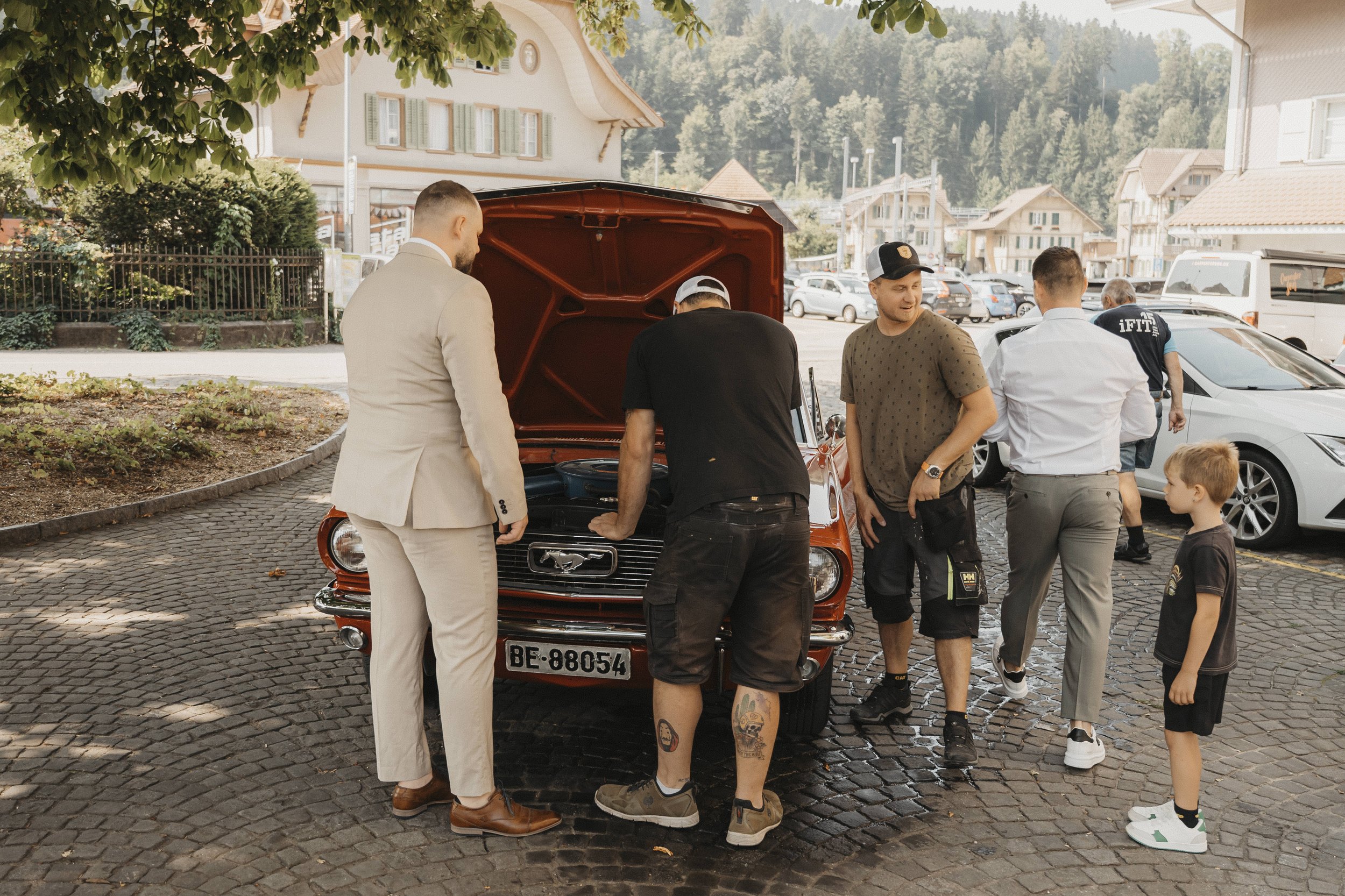 Gruppe von Menschen um eine geöffnete Motorhaube eines roten Classic Ford Mustang Autos herum, auf einem gepflasterten Parkplatz in einem malerischen Dorf mit Hügeln im Hintergrund, bei Tageslicht.