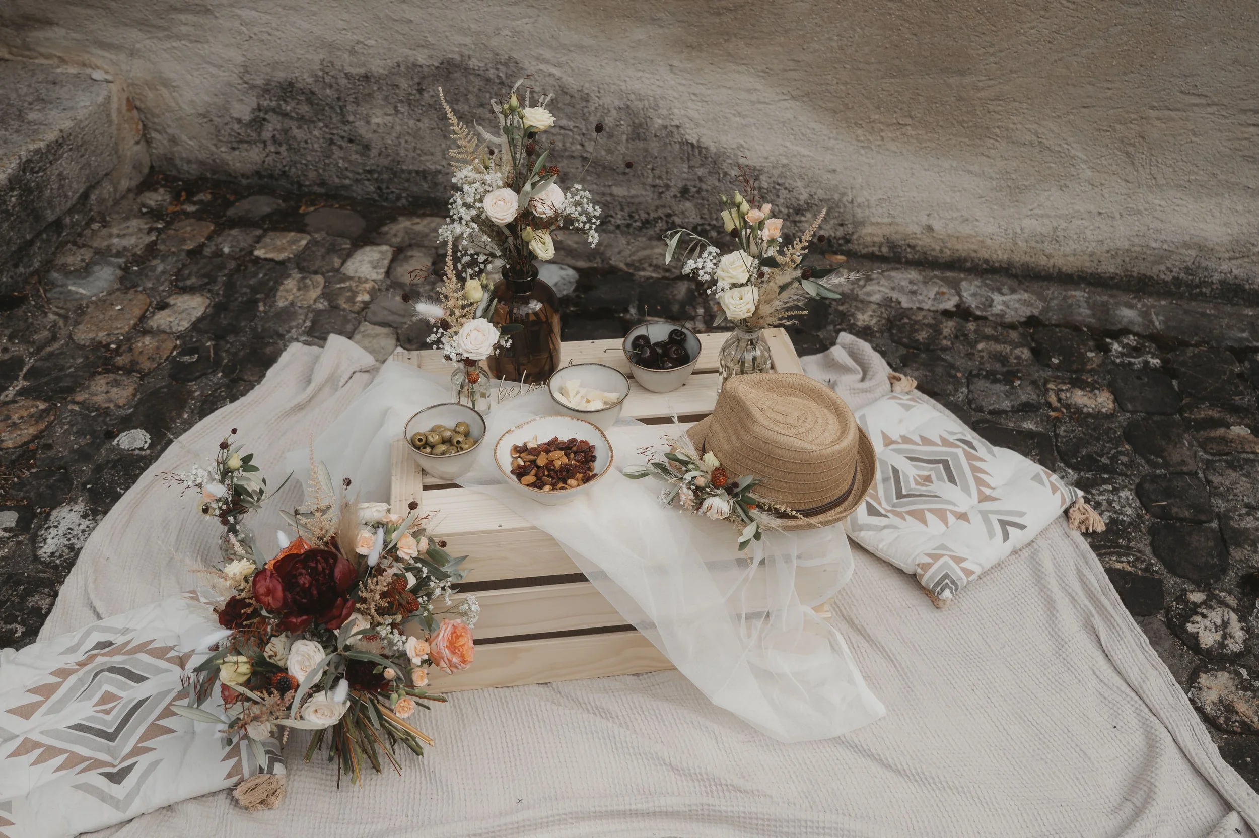 Picknick-Setup auf dem Boden mit Kissen, einem Holztablett, Blumenvasen, einer Hut, Obst und Snacks auf einer Decke