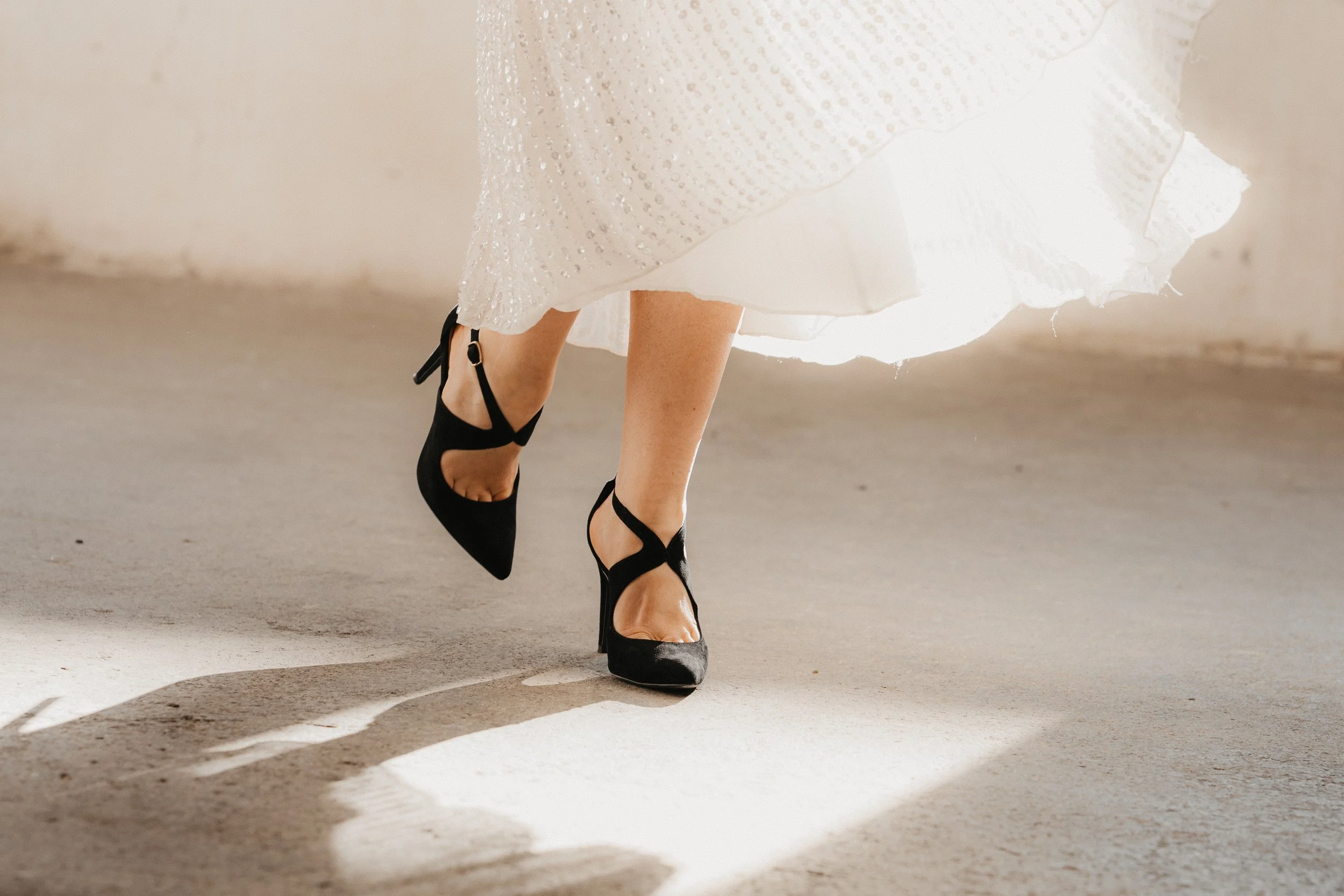 Nahaufnahme von eleganten schwarzen High-Heels an den Füßen einer Person, die ein weißes, schimmerndes Kleid trägt.