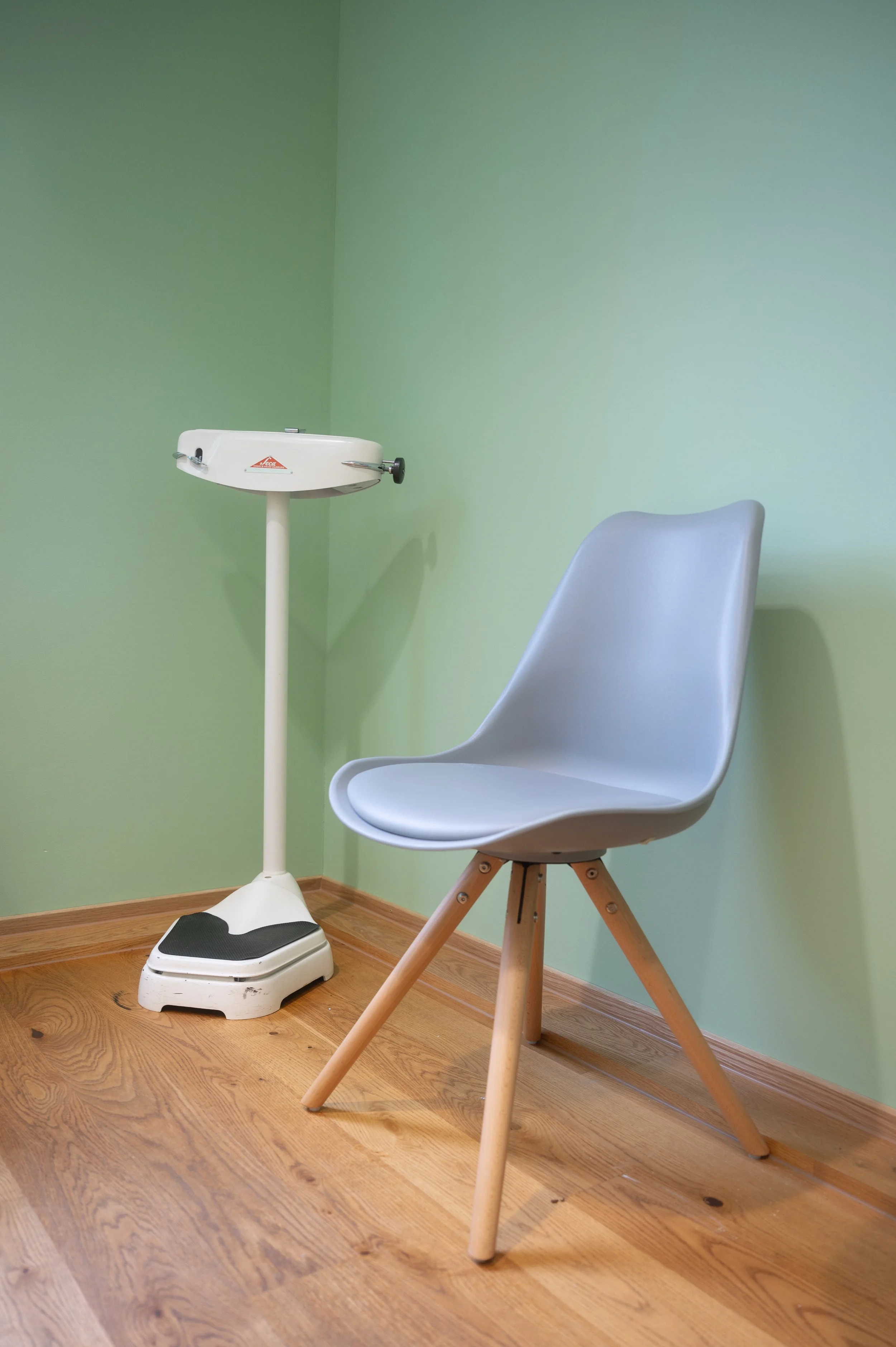 Ein medizinischer Untersuchungsraum mit einer weißen Waage und einem modernen weißen Stuhl vor einer hellgrünen Wand auf Holzboden.