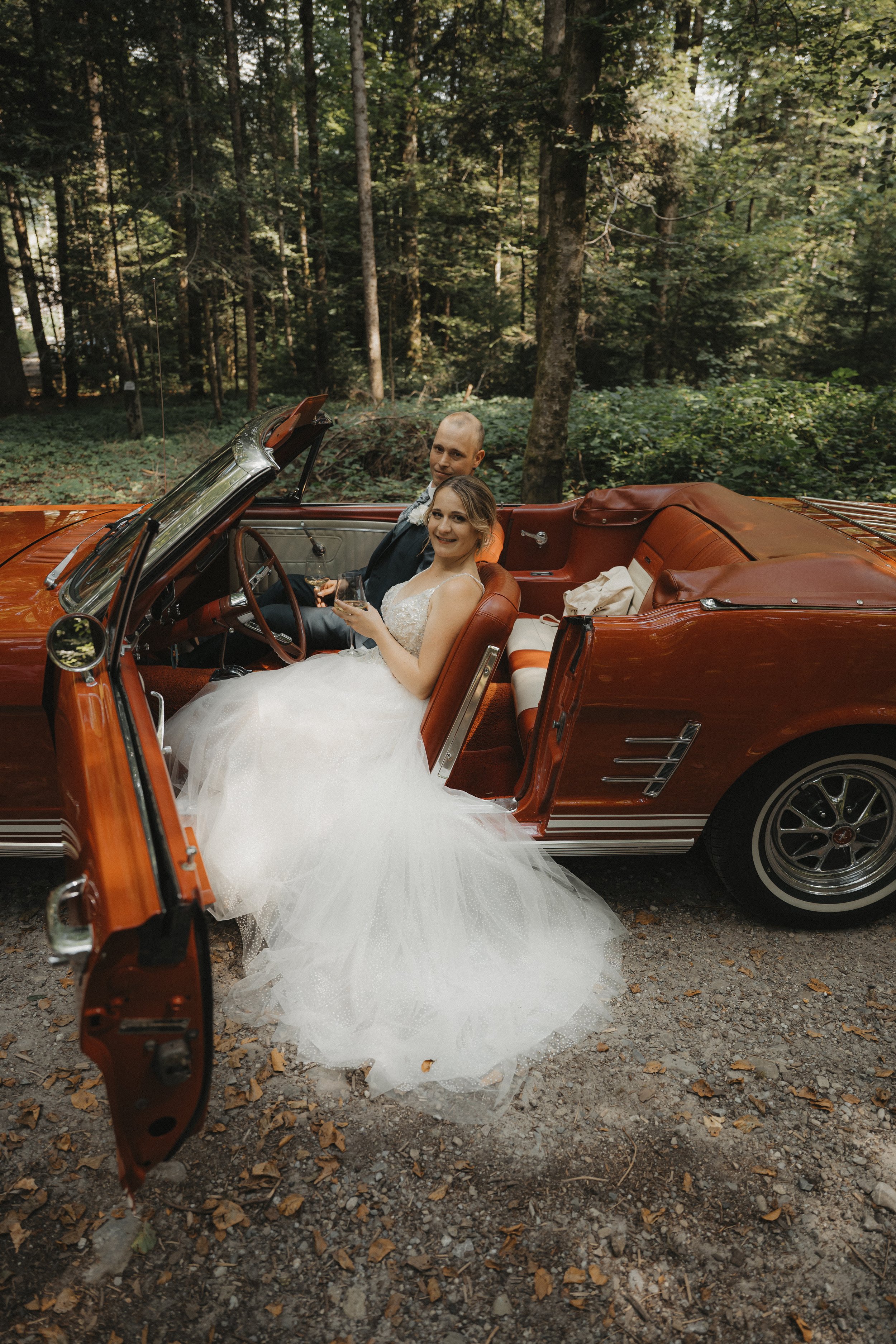 Ein frisch vermähltes Paar sitzt in einem roten Oldtimer inmitten eines Waldes, die Braut trägt ein weißes Hochzeitskleid, beide halten Gläser mit Wein, feiern im Grünen.