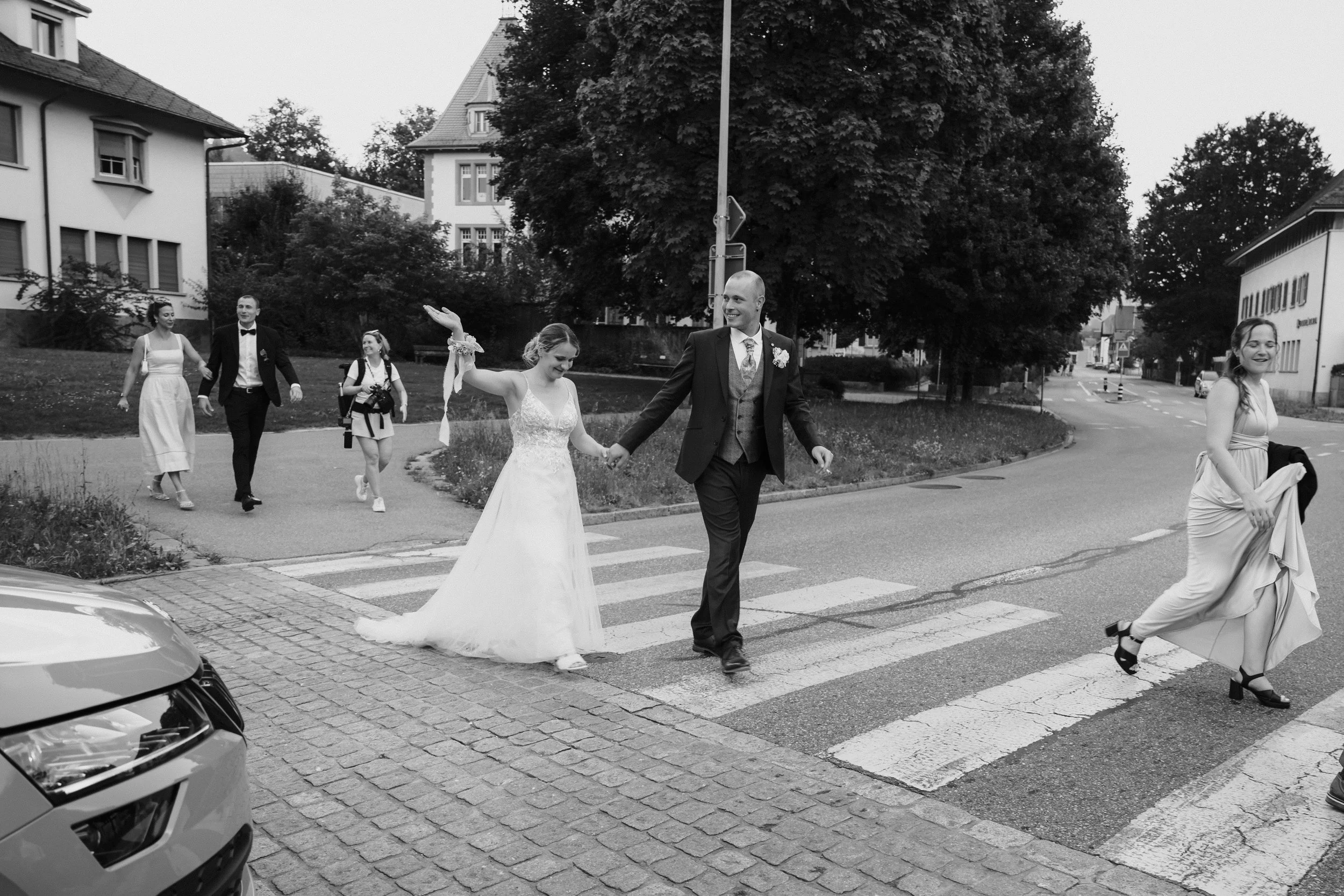 Braut und Bräutigam überqueren einen Zebrastreifen, umgeben von Gästen bei einer Hochzeit im Freien in einer kleinen Stadt, im schwarzen und weißen Foto.