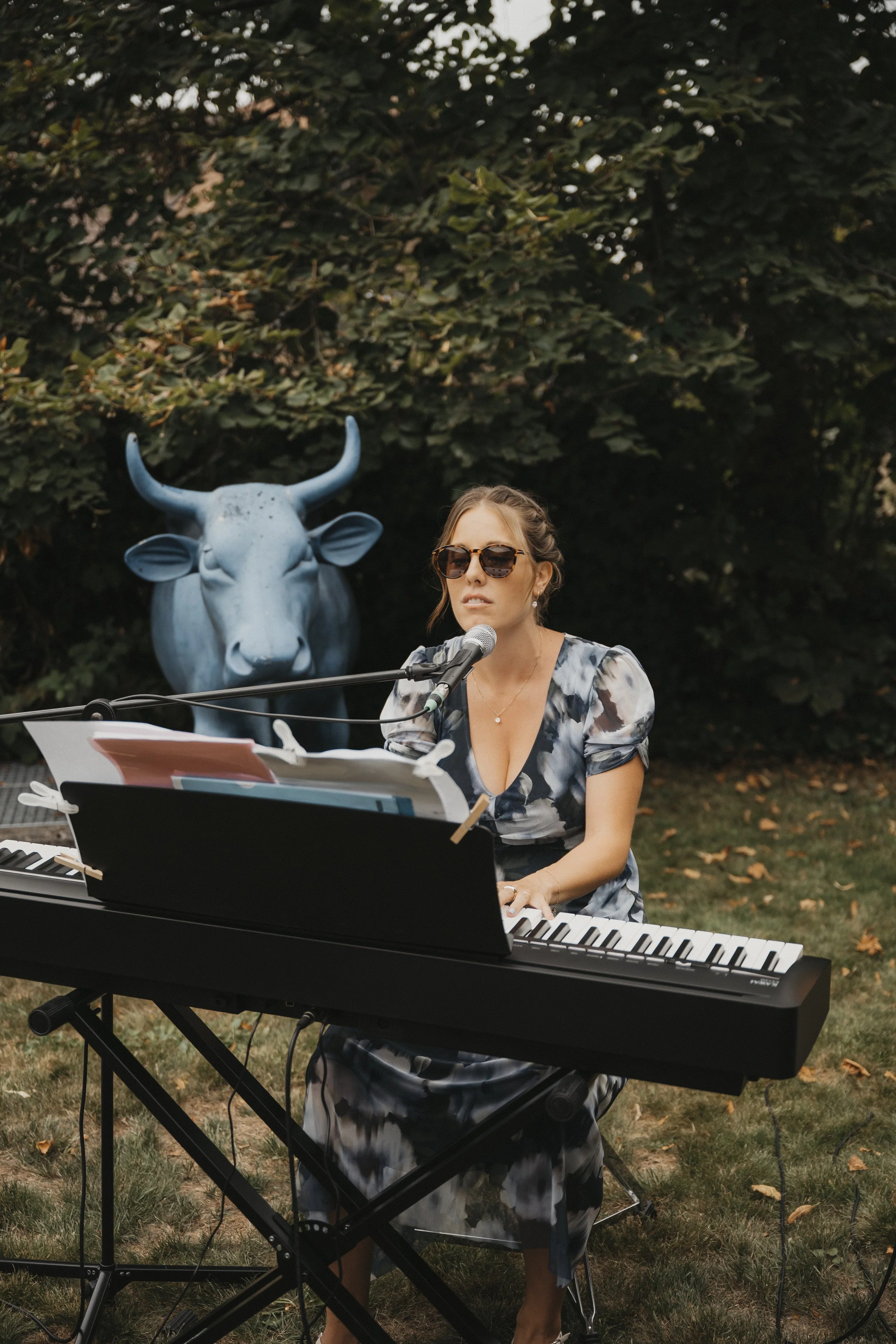 Eine Frau spielt Klavier und singt in einem Garten, im Hintergrund steht eine blaue Kuh-Skulptur aus Metall.