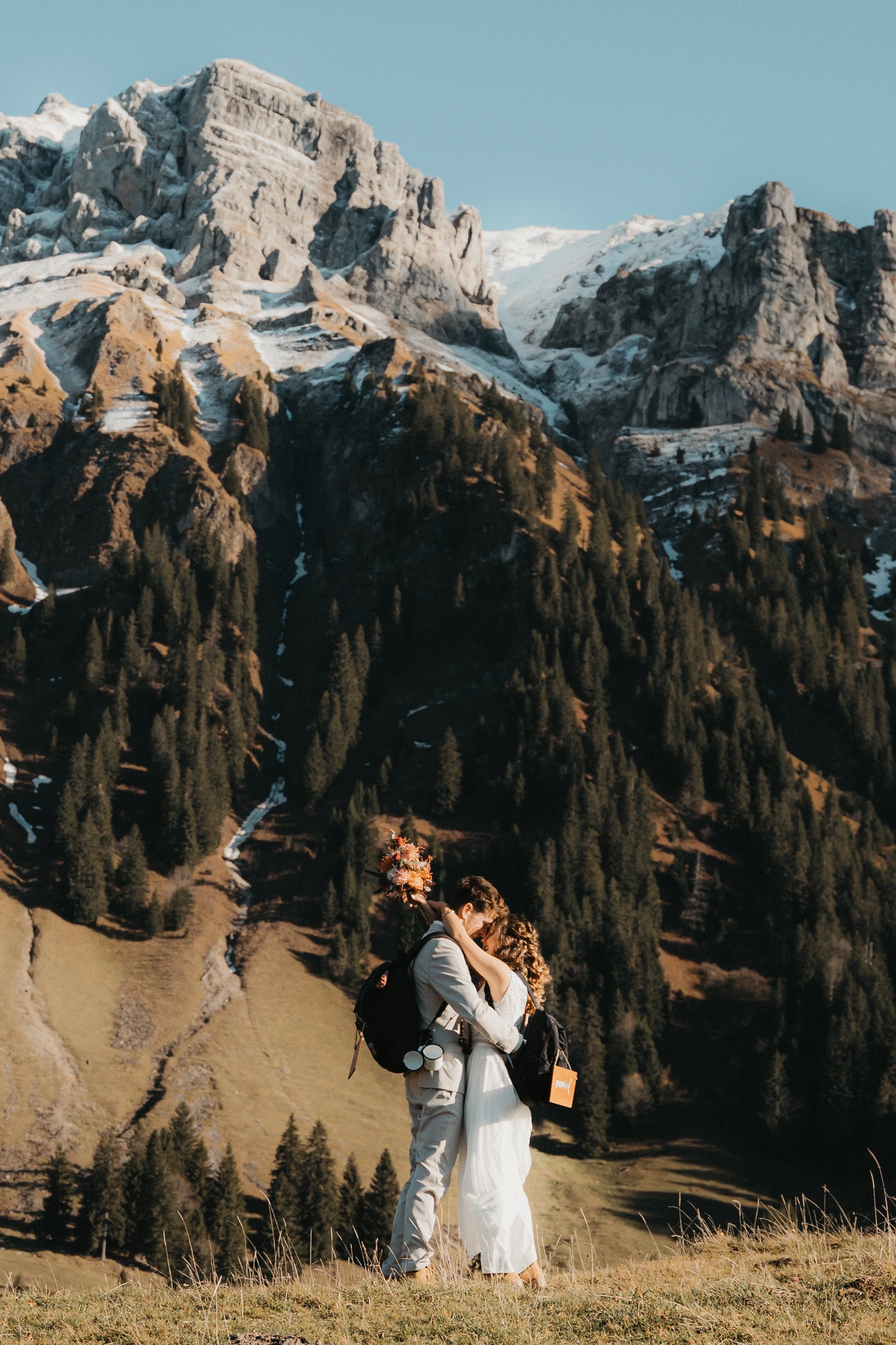 Ein Paar umarmt sich in einer bergigen Landschaft mit hohen, schneebedeckten Bergen und grünen Tannenbäumen im Hintergrund.