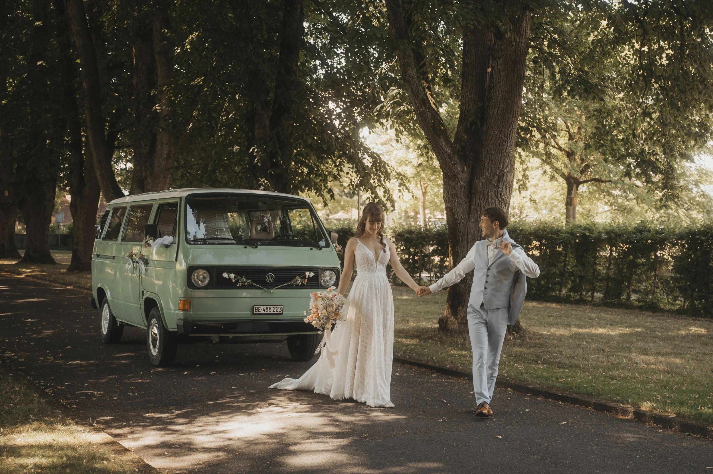 Ein Brautpaar geht Hand in Hand vor einem grünen VW-Bulli in einem parkähnlichen Gebiet mit Bäumen.