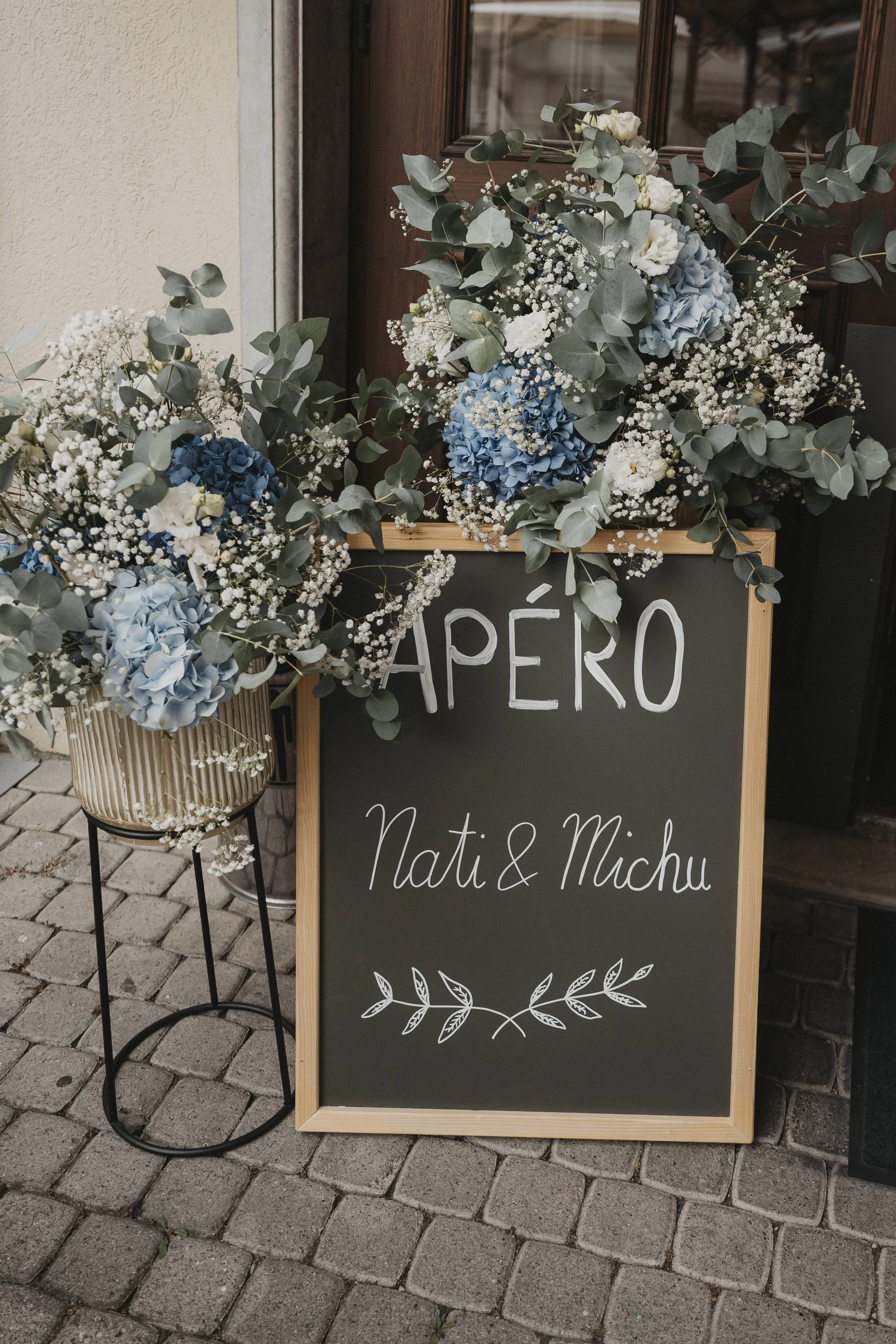 Blumenarrangement mit blauen Hortensien und weißen Blumen, an einem schwarzen Tafel mit Holzrahmen, mit der Aufschrift "HAPÉRO, Nati & Michu", vor einer Steinpflasterstraße.