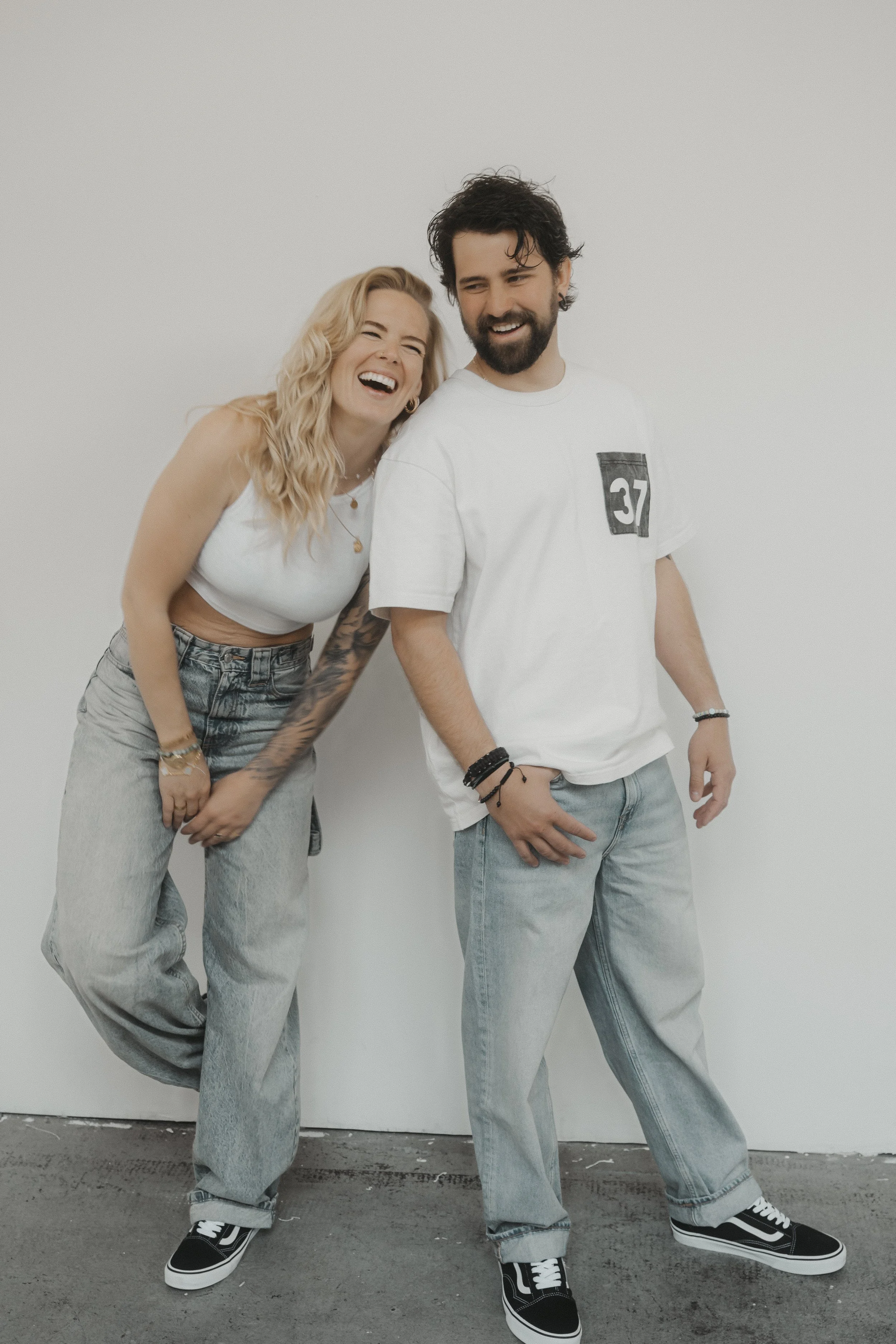 Zwei junge Menschen lachen und posieren vor einer weißen Wand, der eine trägt eine weiße Weste und die andere ein weißes T-Shirt mit der Zahl 37, beide in Jeans und Turnschuhen.