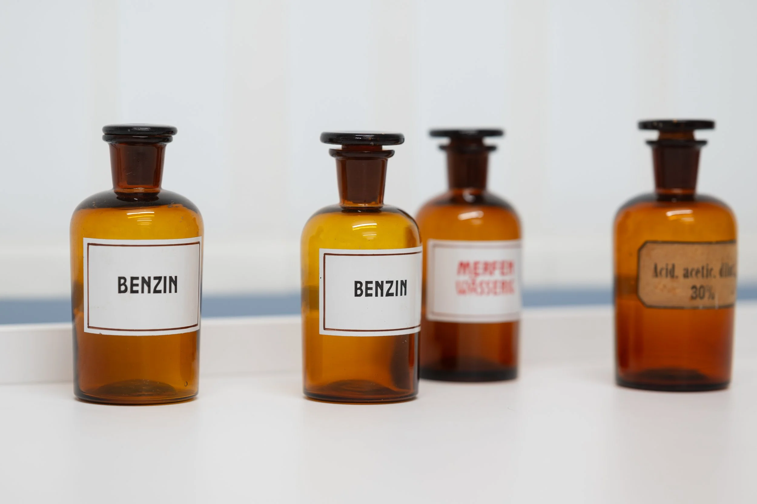 Vier braune Glasflaschen mit weißen Etiketten, zwei mit der Aufschrift 'BENZIN', die anderen mit verschiedenen chemischen Bezeichnungen.