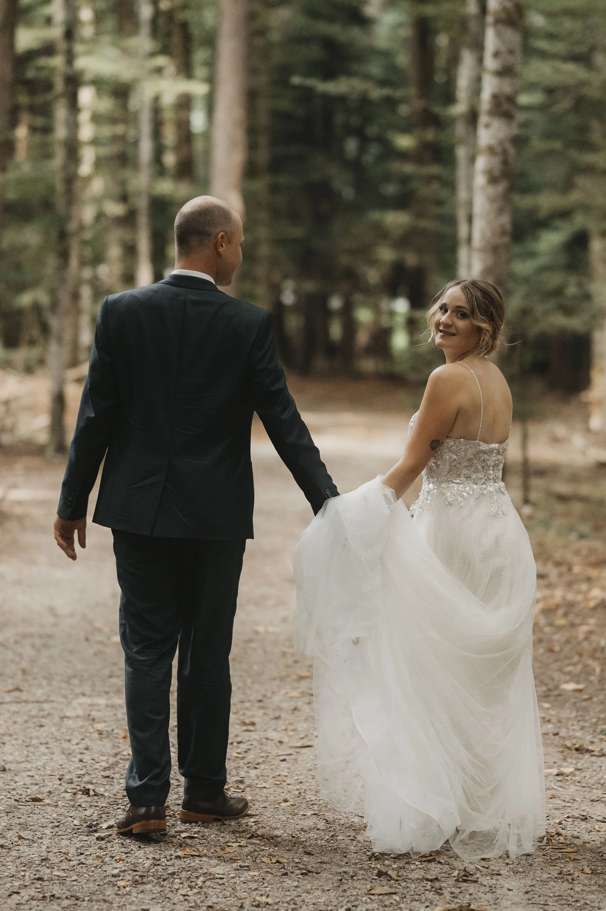 Ein brautpaar geht hand in hand durch einen Wald. Die Braut trägt ein weißes Hochzeitskleid und lächelt, während der Bräutigam einen dunklen Anzug trägt.