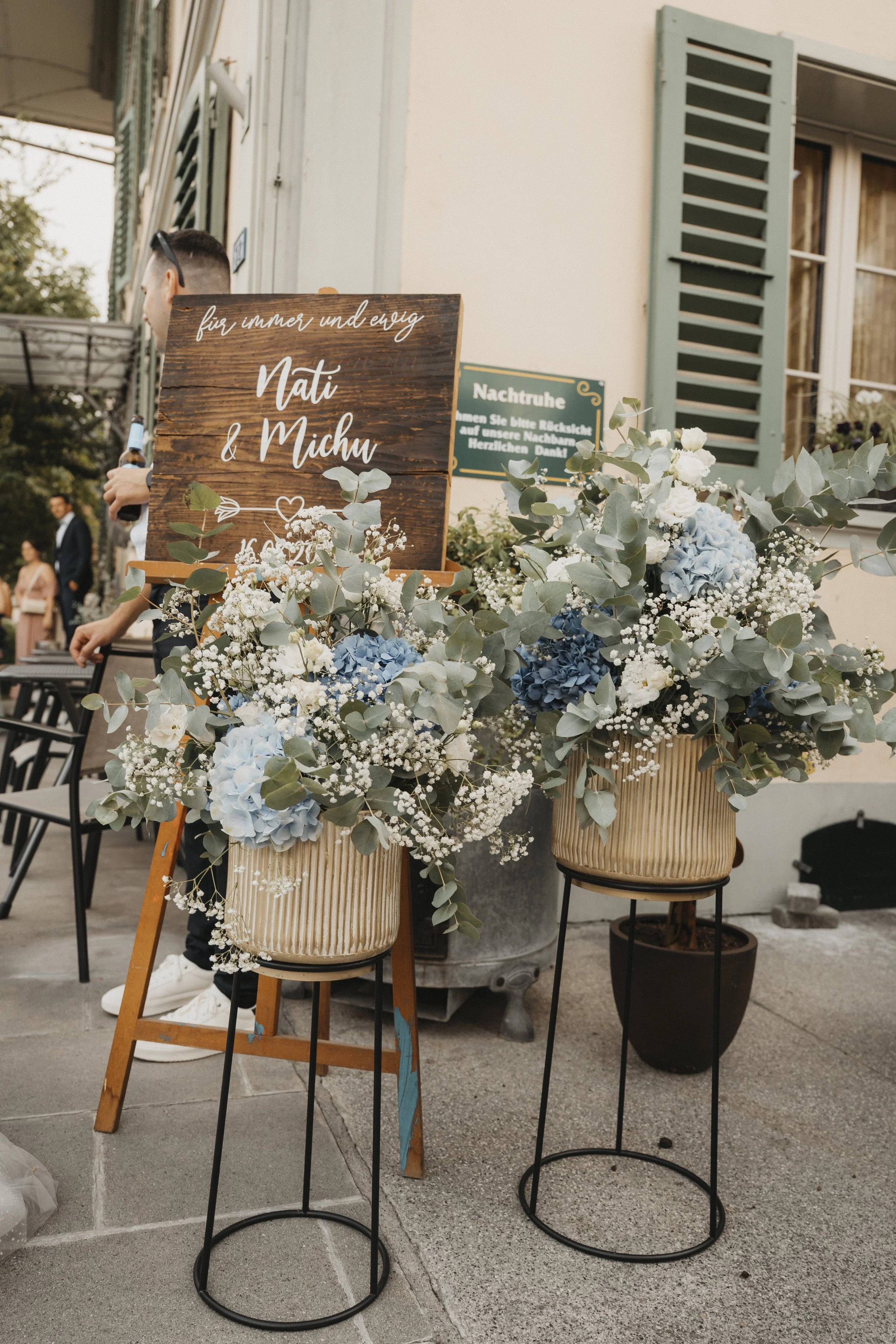 Blumenarrangements mit blauen Hydrangeas und weißen Blumen vor einem Hochzeitsbanner mit den Namen Nati und Michu.