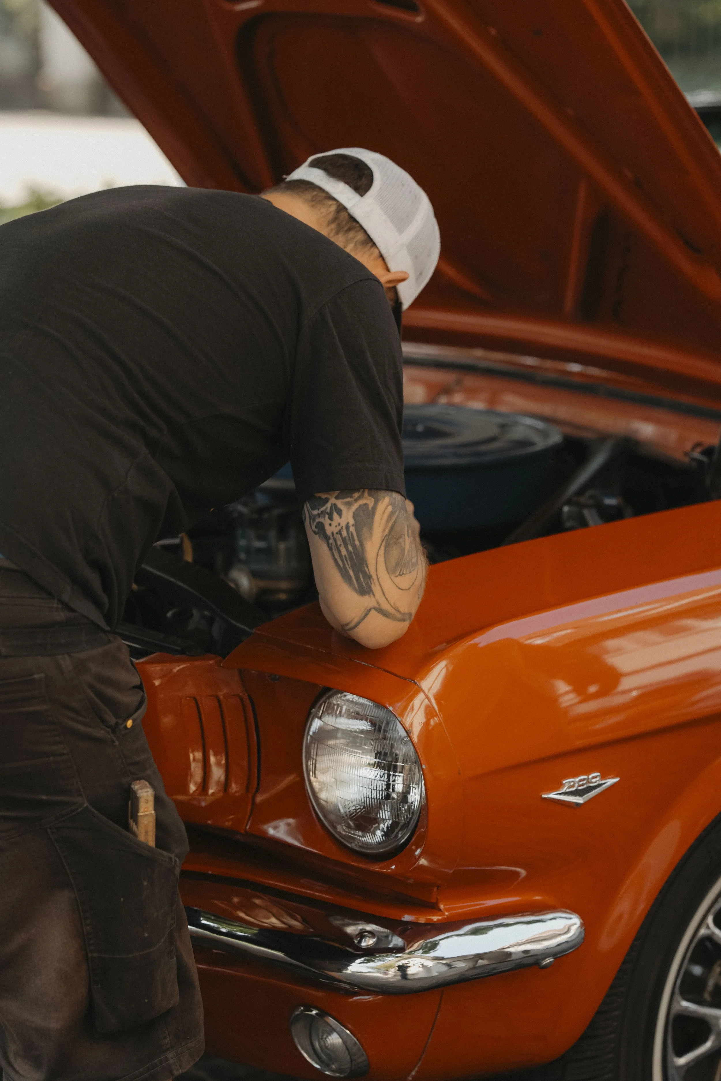 Ein Mann mit Tattoo am Arm arbeitet an einem orangefarbenen Auto mit geöffnetem Motorhaube.