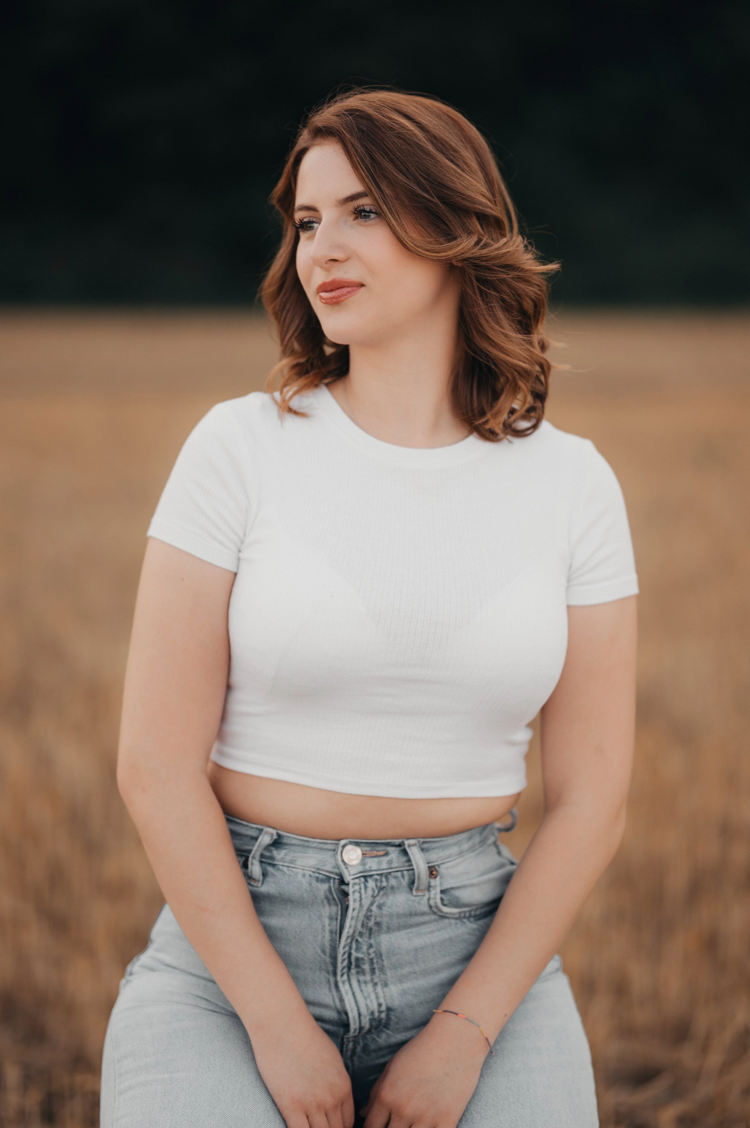Eine junge Frau mit roten, gewellten Haaren trägt ein weißes T-Shirt und Jeans, steht in einer offenen Feldlandschaft bei sonnigem Wetter.