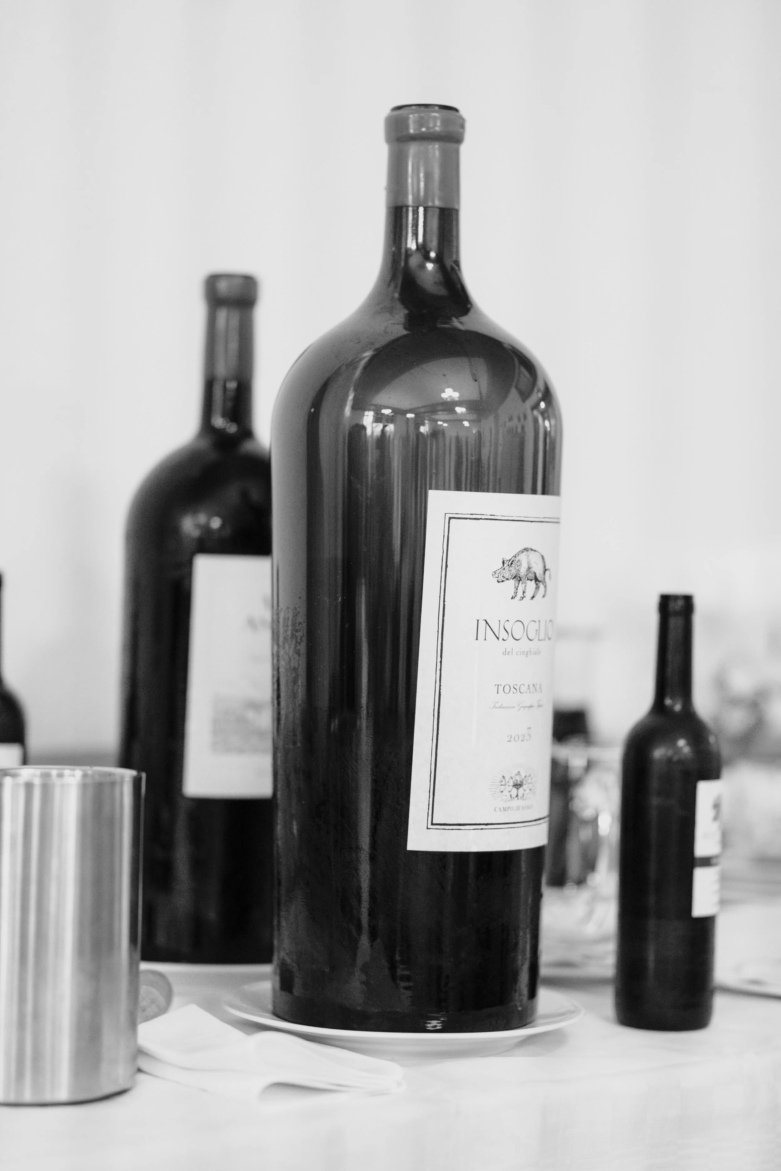 Schwarbe Weinflasche mit Etikett, im Hintergrund weitere Weinflaschen, auf weißem Tuch liegende Papiertaschentücher, und ein metallner Becher.