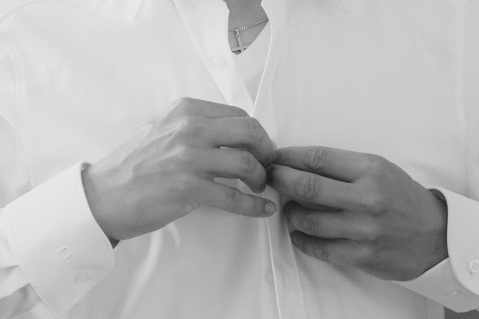 Eine Person in einem weißen Hemd, die anknöpft, mit einer silbernen Halskette mit Kreuz.