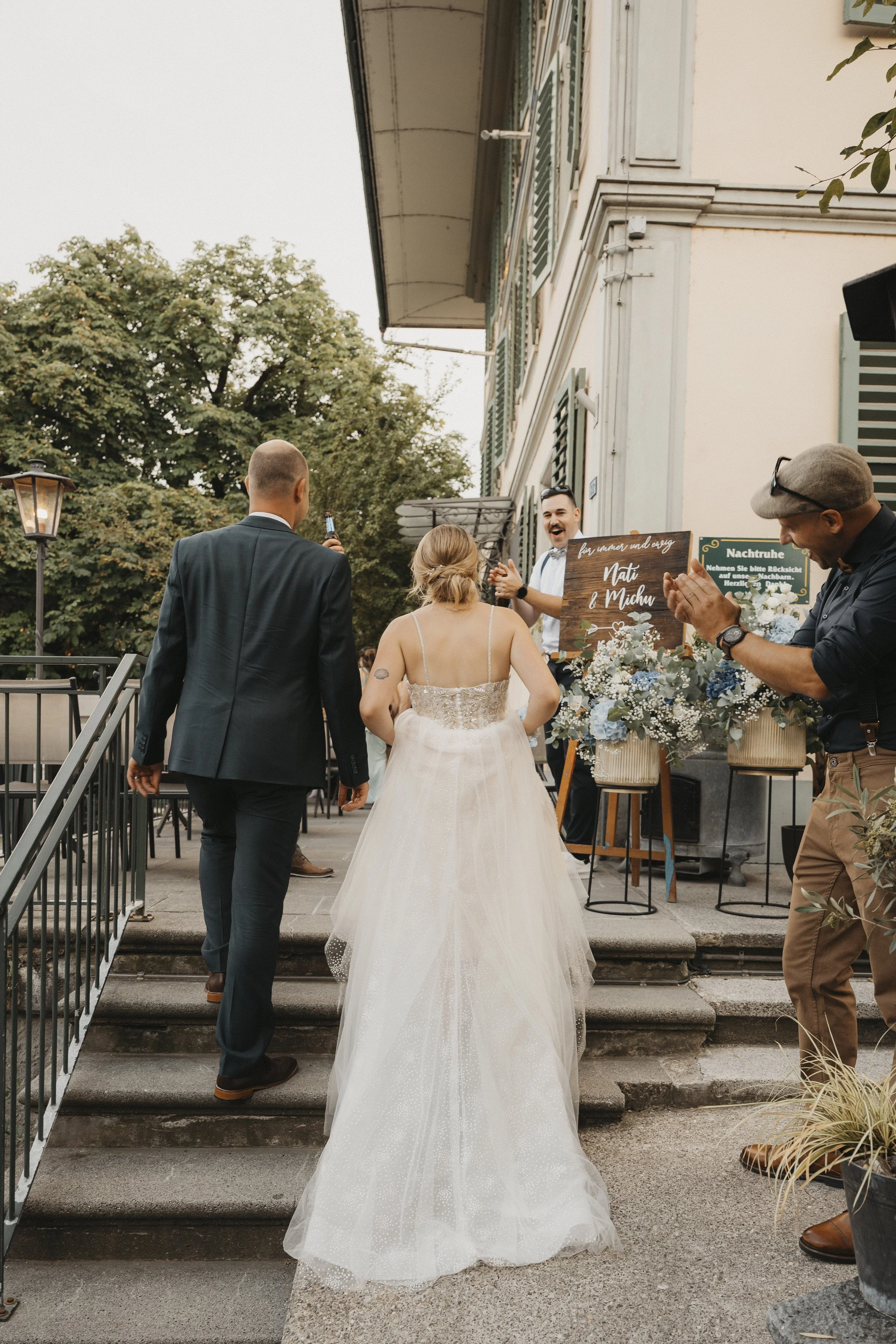 Hochzeitspaar steigt die Treppe hoch, umarmt sich, während Gäste applaudieren und Fotos machen. Im Hintergrund ein Willkommensschild und Blumendekoration.
