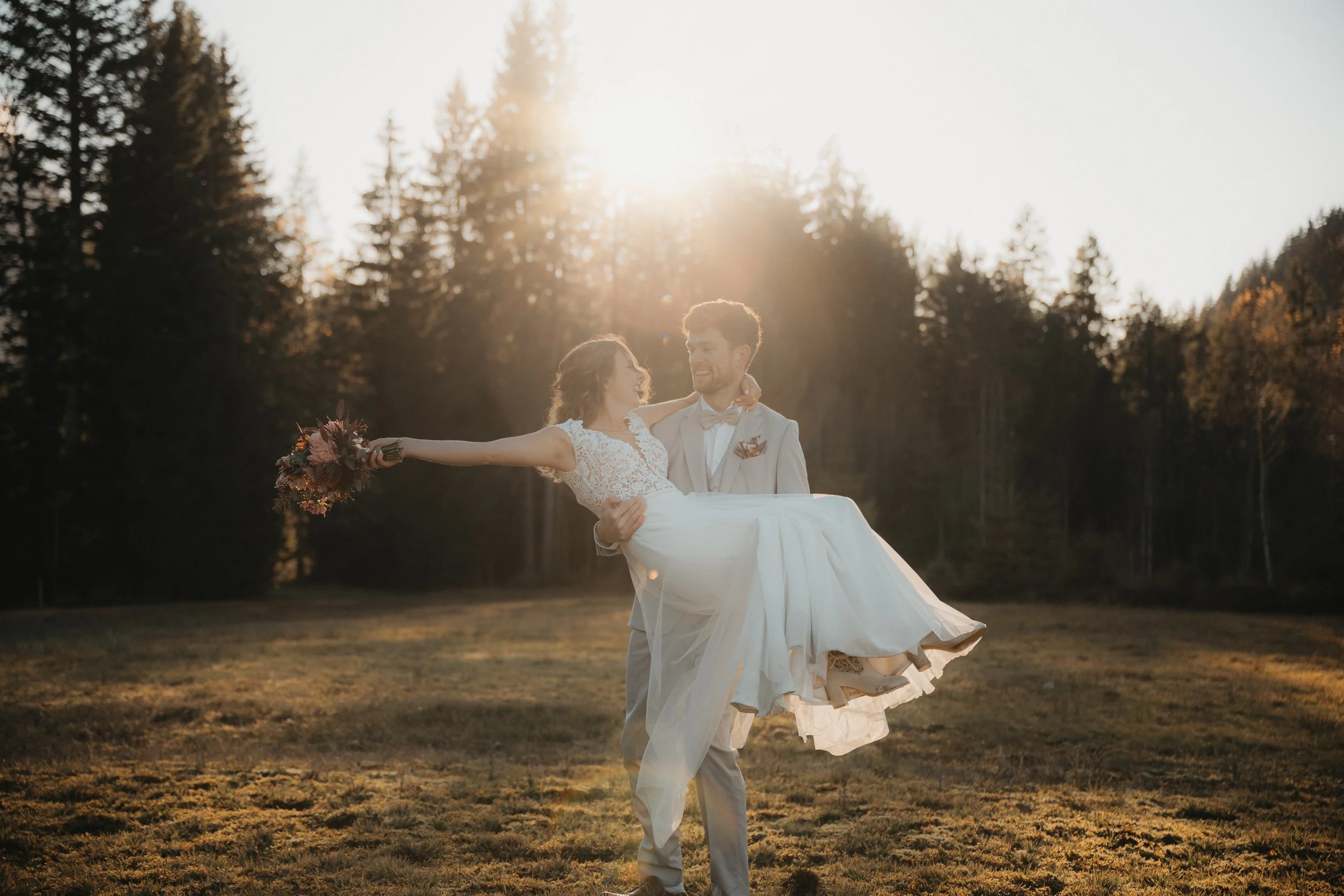 Ein glückliches Brautpaar im Sonnenuntergang, der Bräutigam trägt einen hellen Anzug und trägt die Braut, die ein weißes Hochzeitskleid trägt, in den Armen bei einem Spaziergang im Freien auf einer Wiese mit Bäumen im Hintergrund.