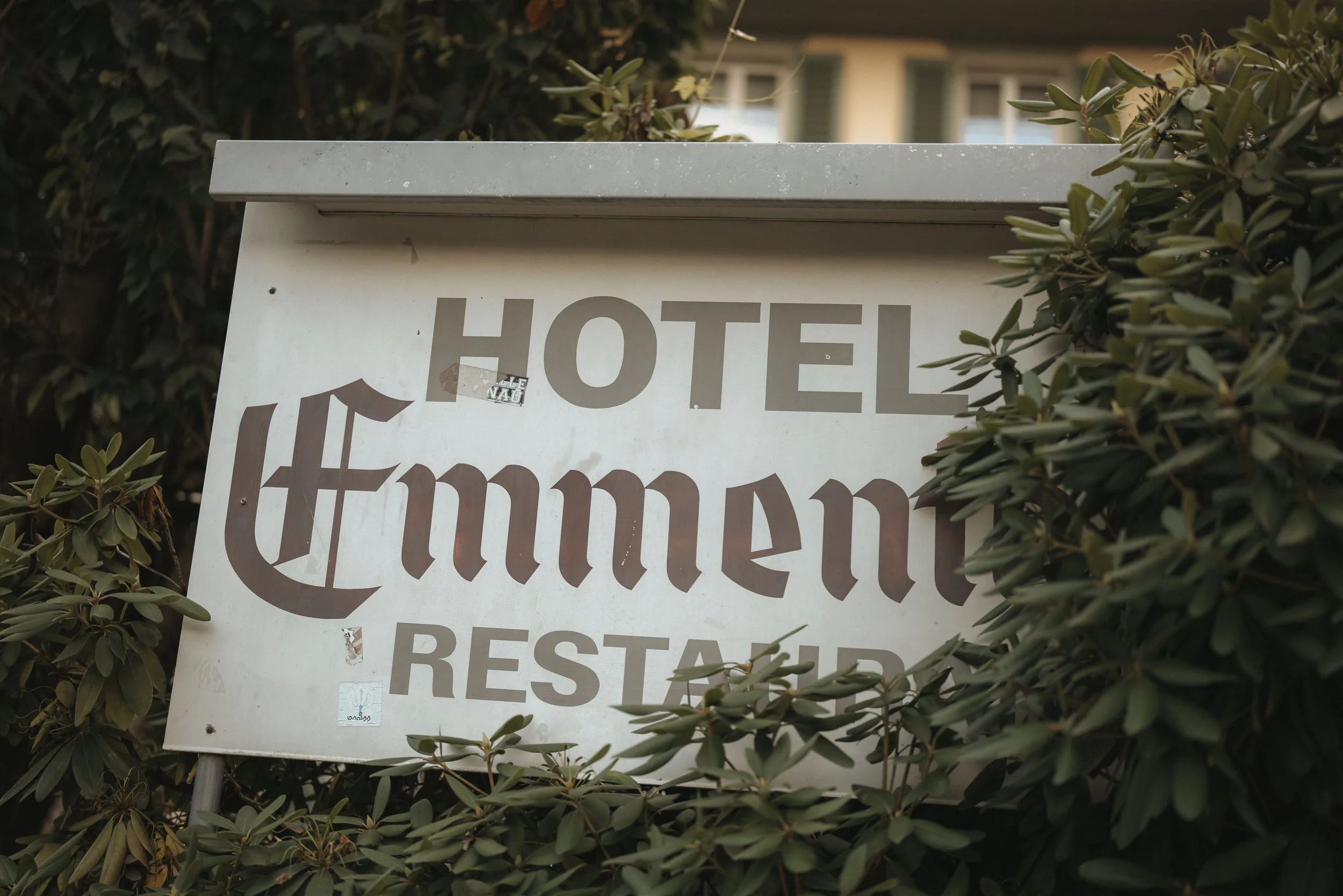Ein Hotelschild mit dem Namen 'Himmel' in einer Vegetation, es ist leicht beschädigt und hängt vor einem Gebäude.