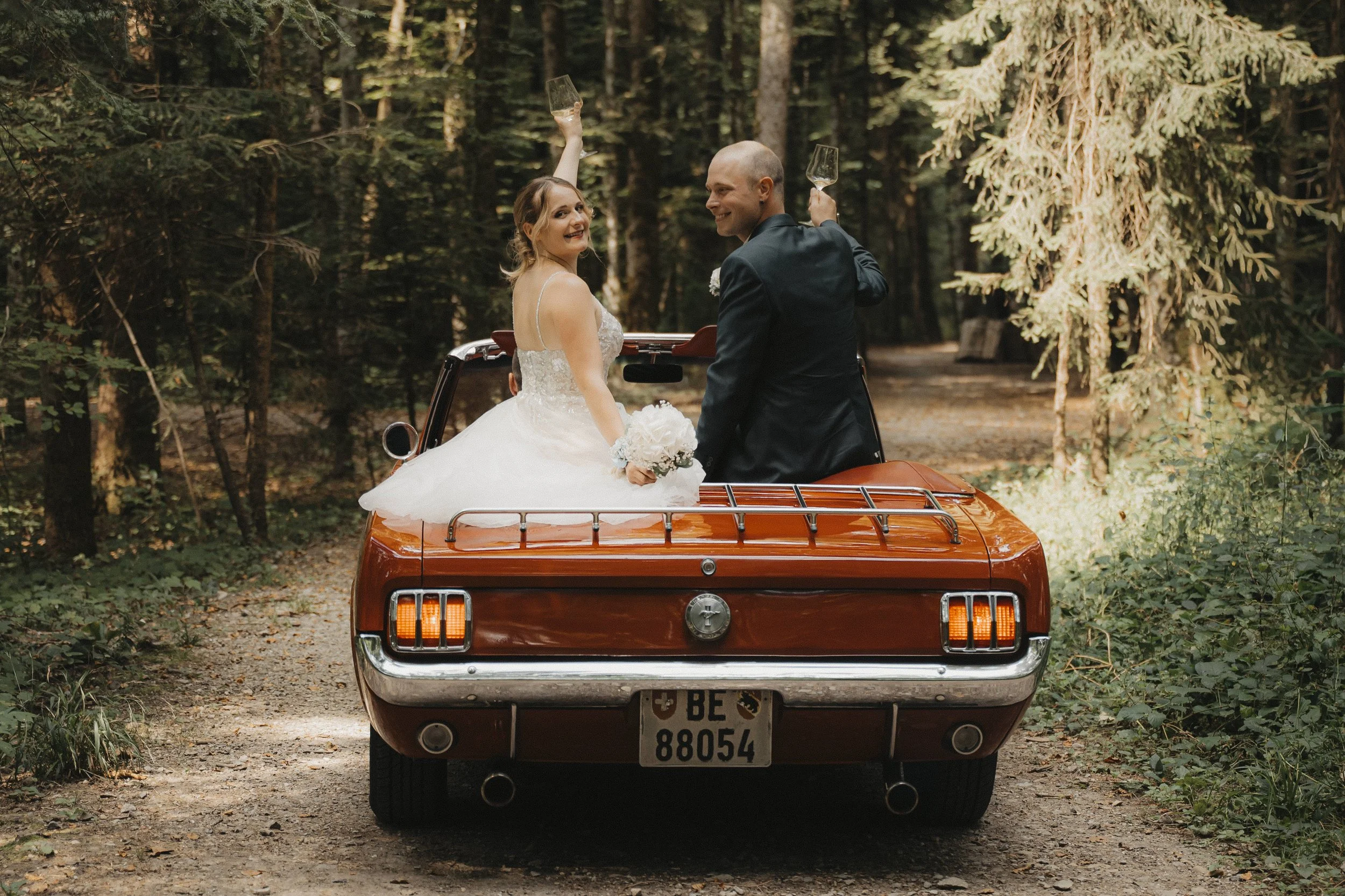 Ein Brautpaar sitzt auf der Ladefläche eines roten Oldtimers, umgeben von einem Wald. Die Braut trägt ein weißes Hochzeitskleid und hält einen Brautstrauß, während sie lächelnd in die Kamera schaut. Der Bräutigam trägt einen schwarzen Anzug und hält 