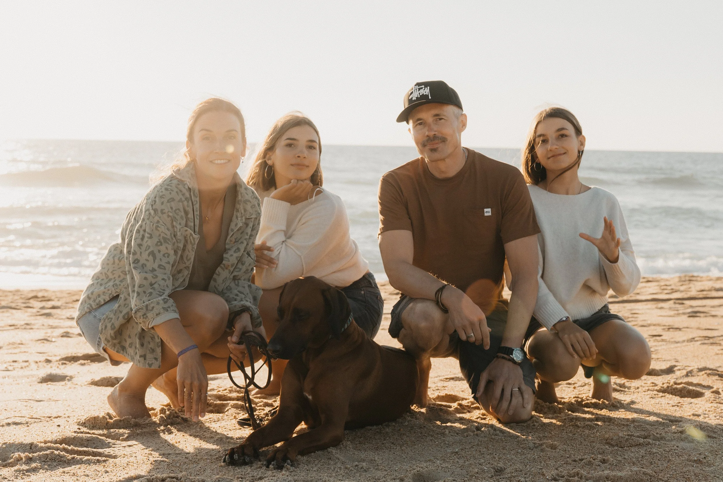 Vier Menschen und ein Hund am Strand bei Sonnenuntergang, im Hintergrund das Meer.