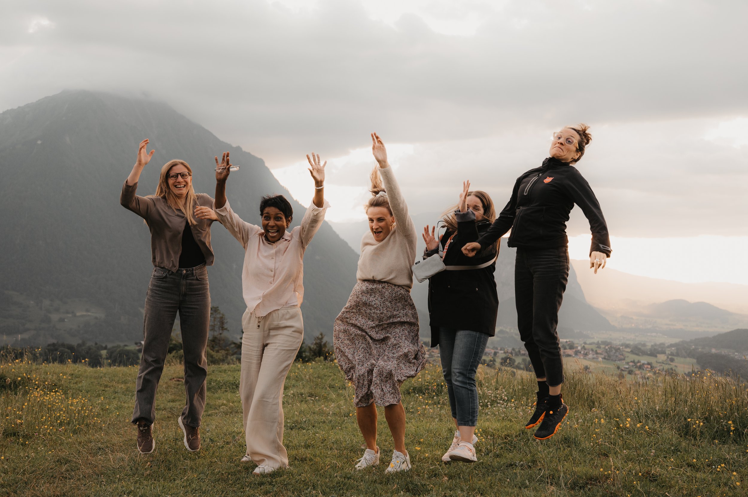 Fünf Frauen springen in einem grünen Hügel mit Bergen im Hintergrund bei Sonnenuntergang und lachen.